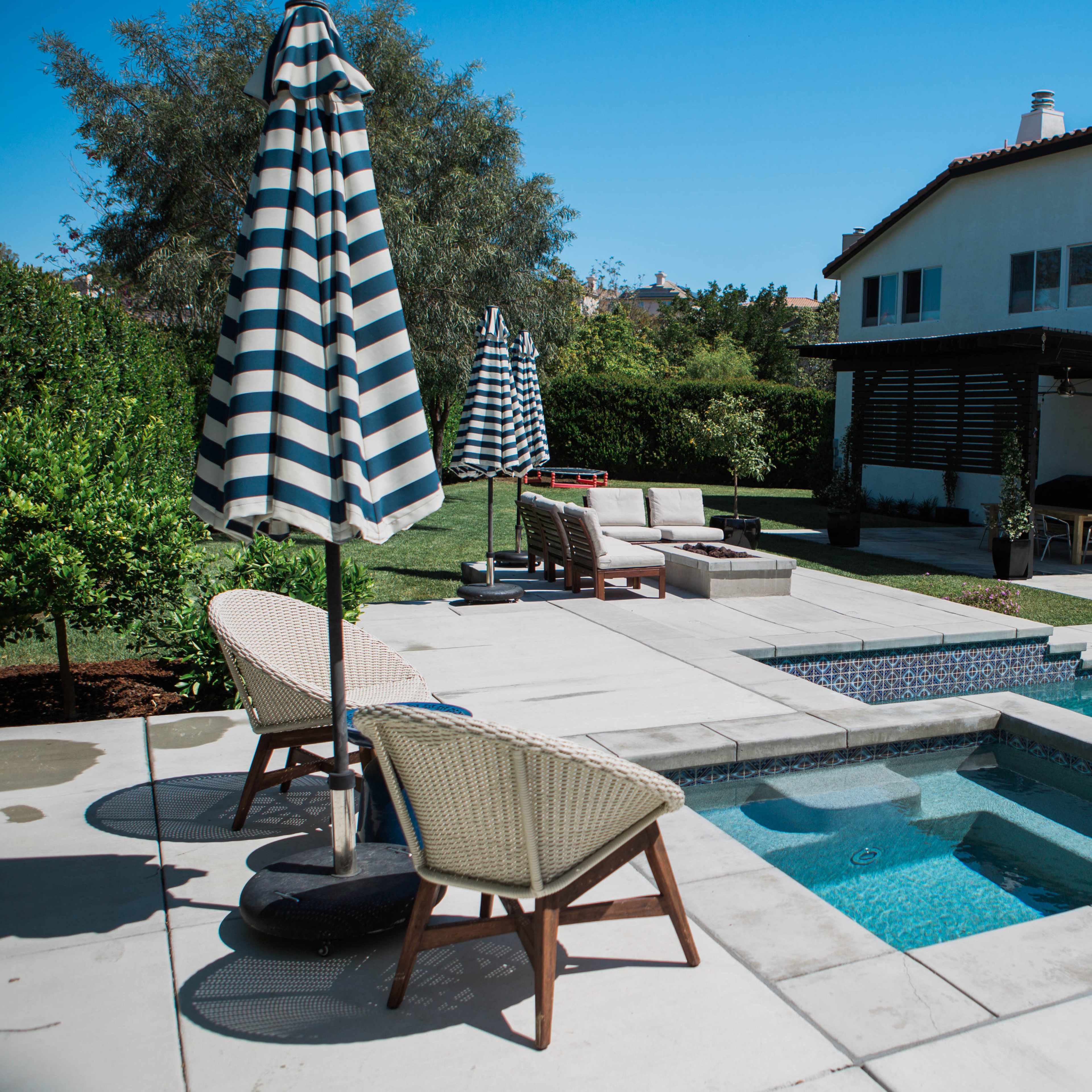 A residential backyard featuring a swimming pool, a spa, lounge chairs, and striped umbrellas beside a two-story white house.