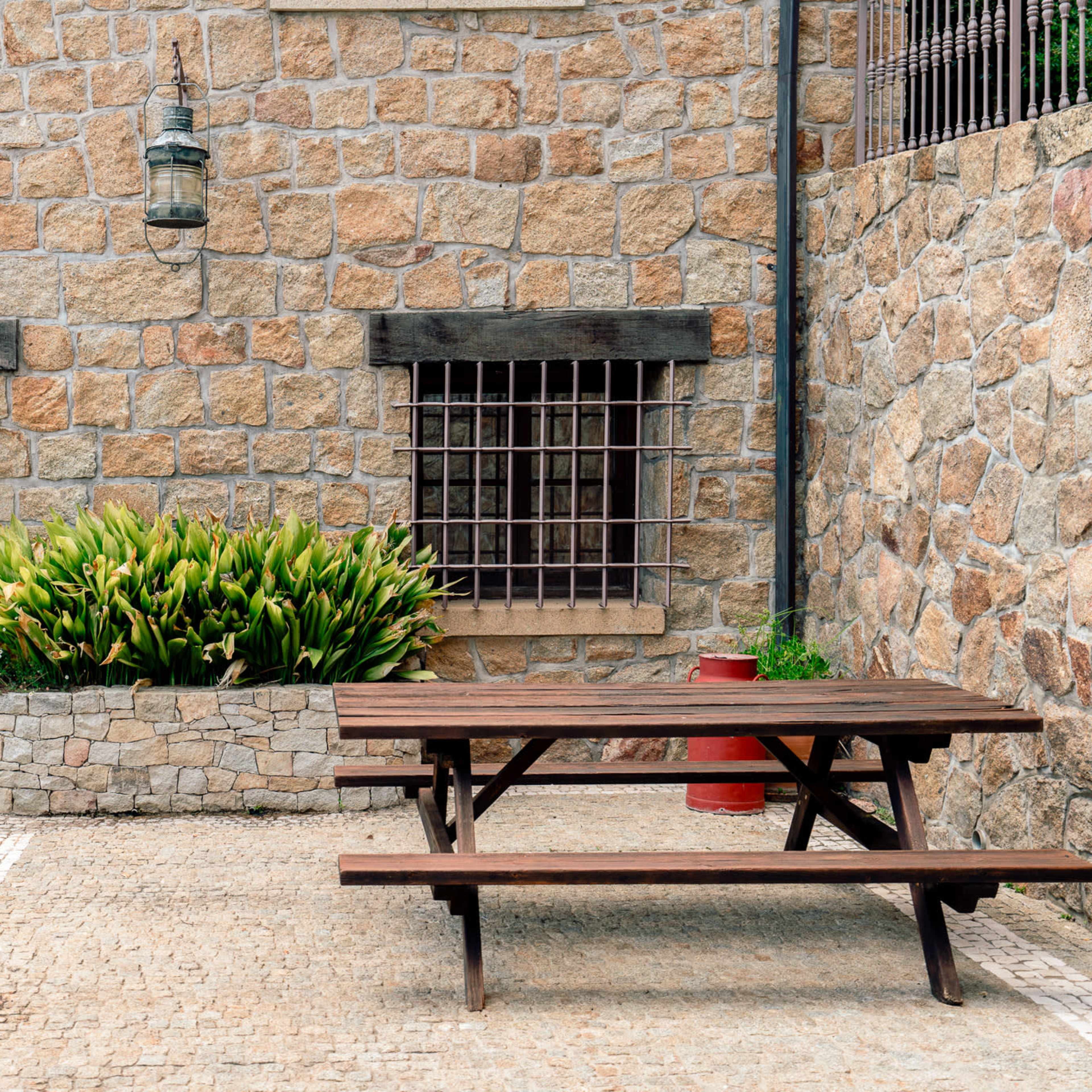 A stone wall features two barred windows and a wooden picnic table in a paved courtyard surrounded by greenery.