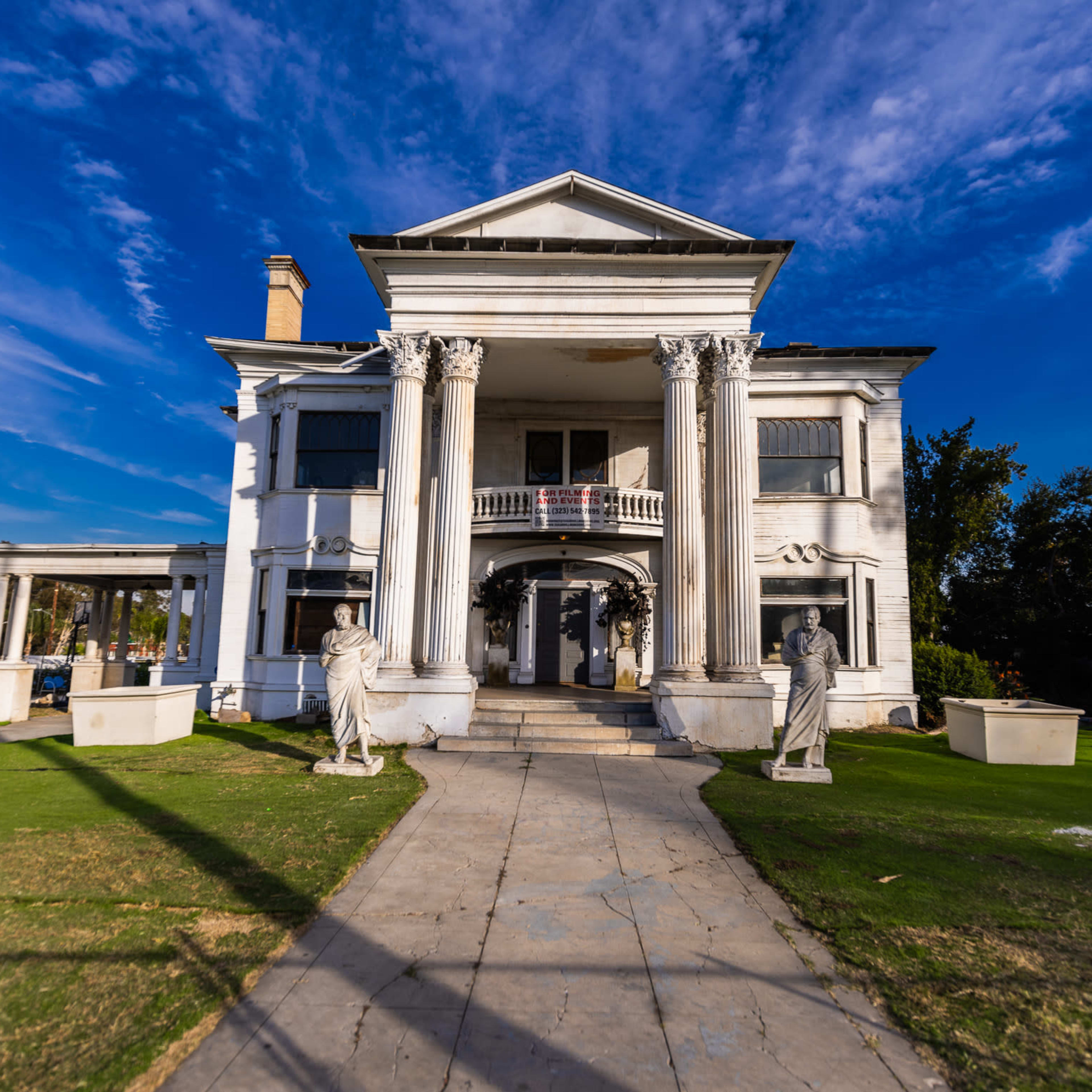 A large, white, classical-style mansion with tall columns and two statues flanking the entrance, set against a clear blue sky.