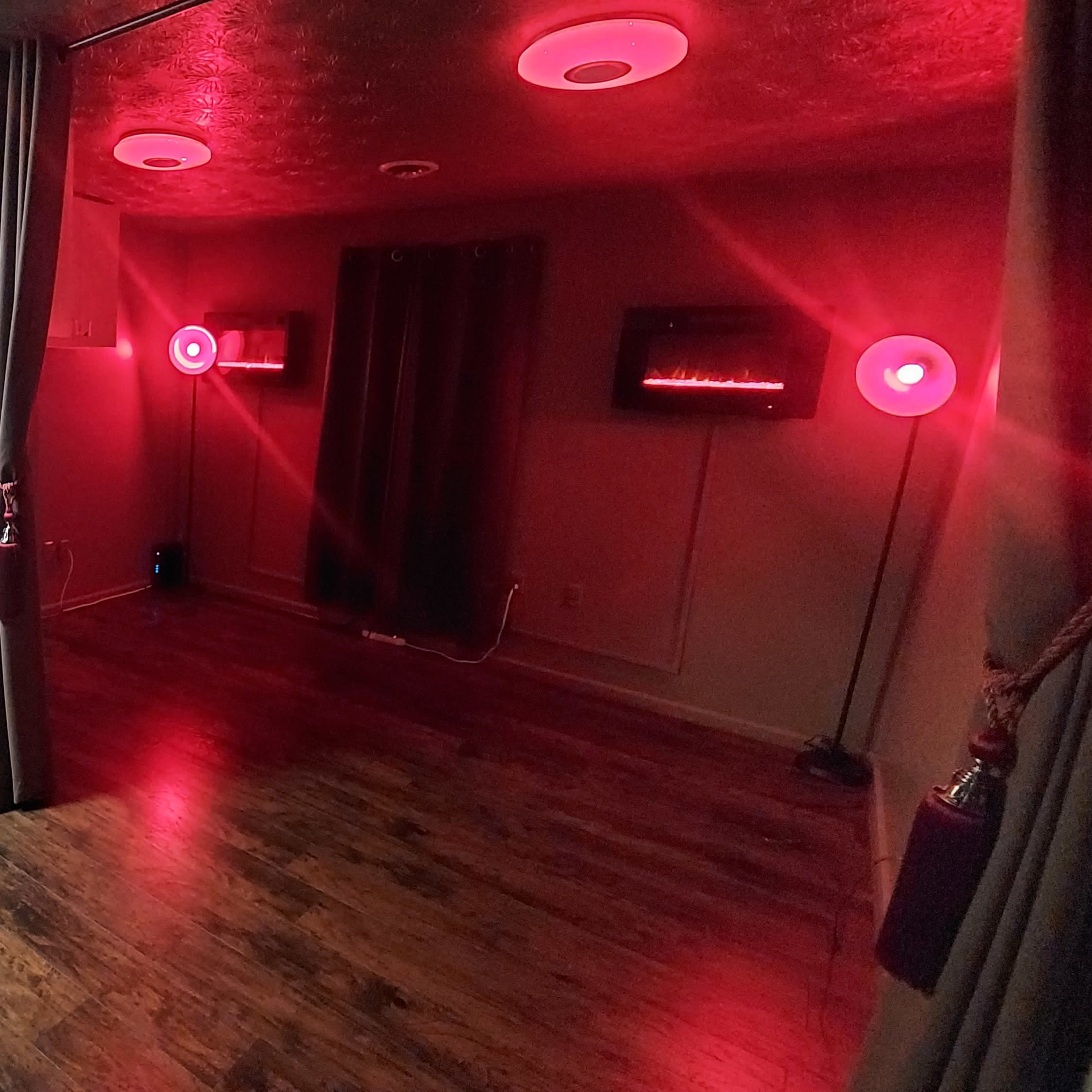 A dimly lit room with wooden flooring, red lighting from circular fixtures, and two screens mounted on the walls, flanked by curtains.
