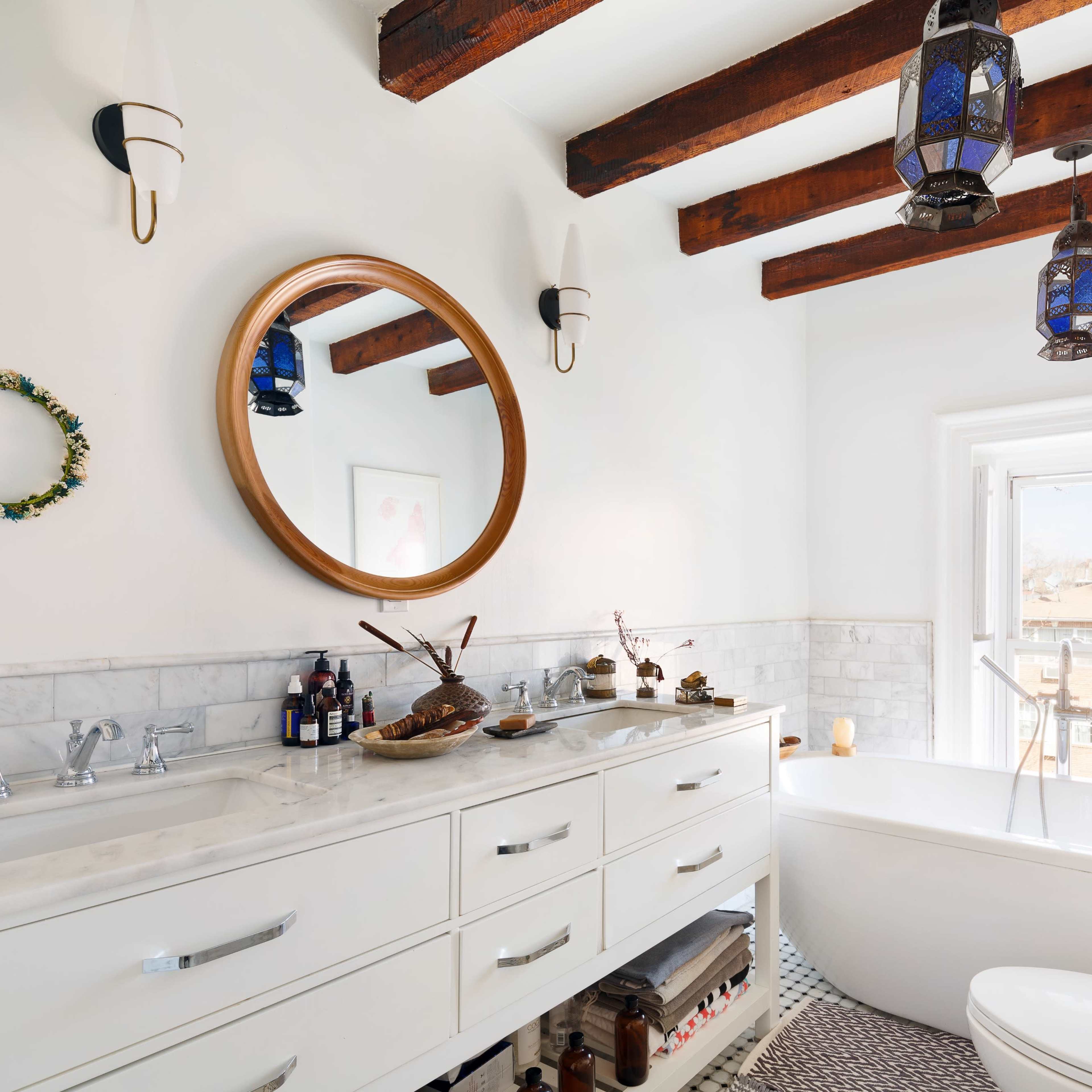 A bright bathroom features a freestanding bathtub, double sink vanity with a round mirror, and pendant lights hanging from wooden beams.