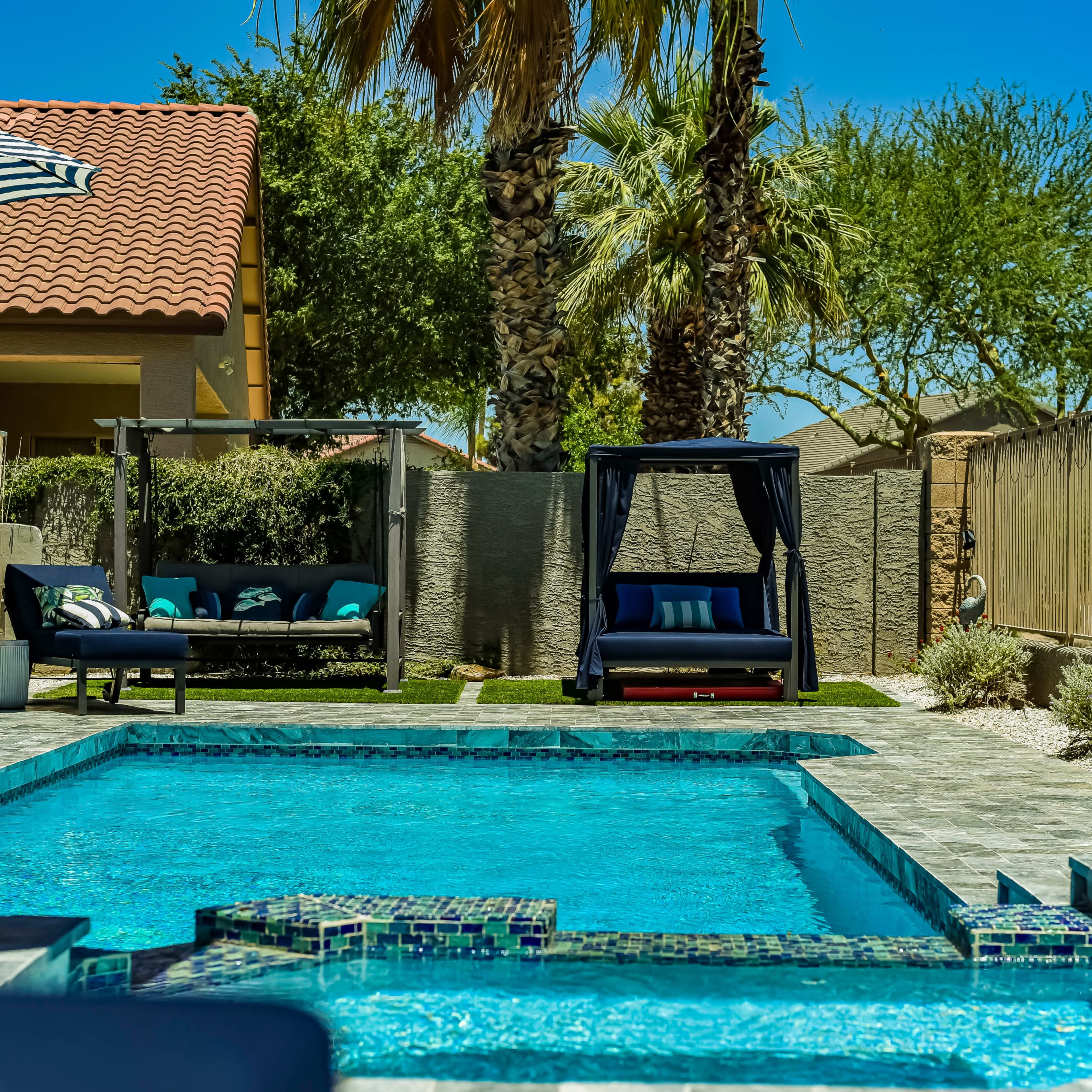 A residential backyard featuring a swimming pool, lounge chairs, a swinging daybed, and palm trees under a clear blue sky.