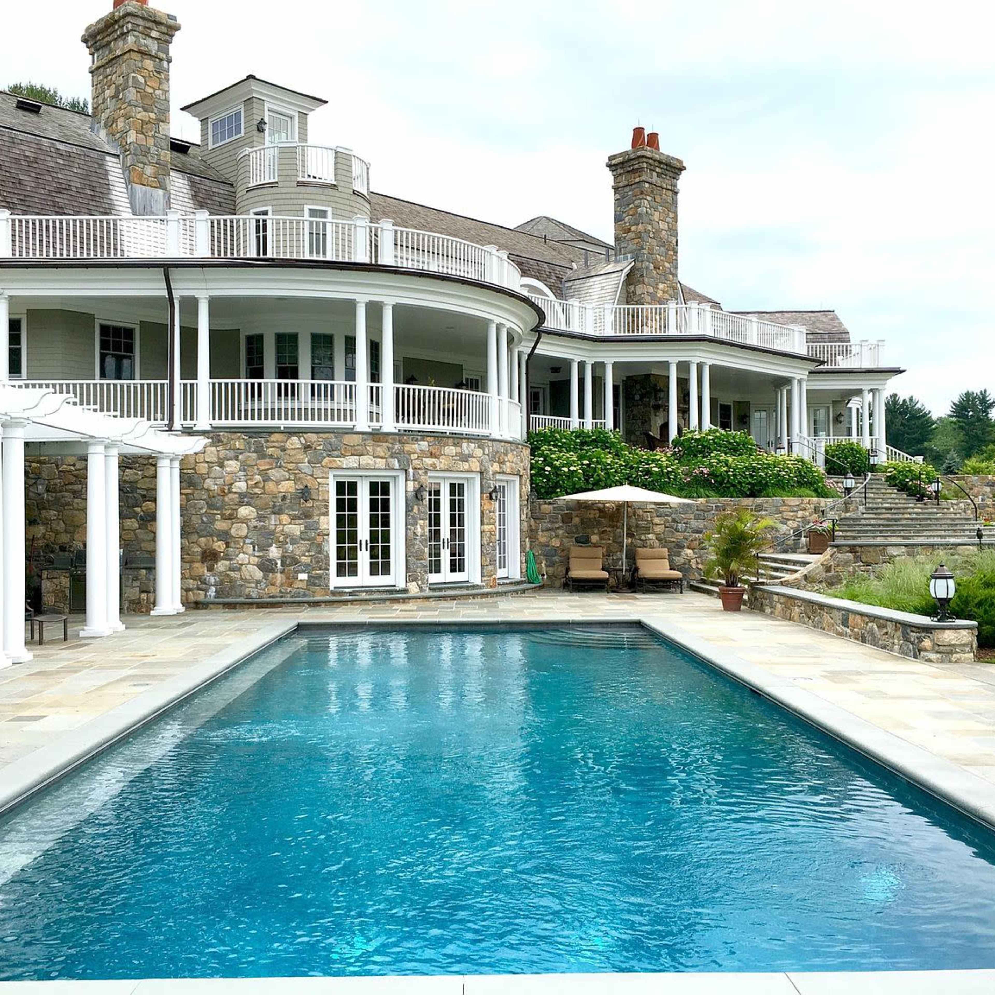 A large stone house with multiple levels and balconies overlooking a rectangular swimming pool surrounded by landscaped gardens.