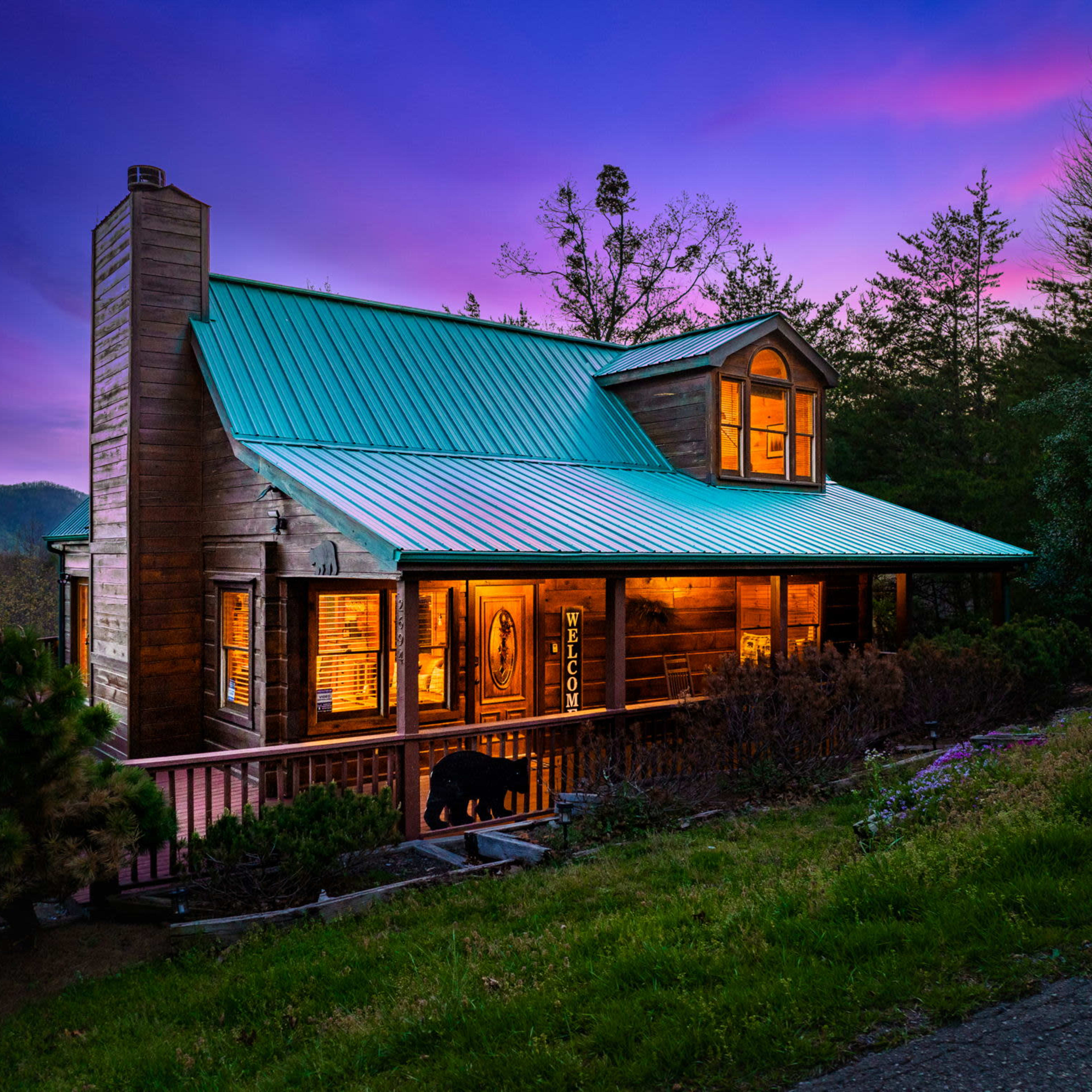 A wooden cabin with a green metal roof, illuminated by warm lights against a purple and blue twilight sky, surrounded by trees and landscaped greenery.