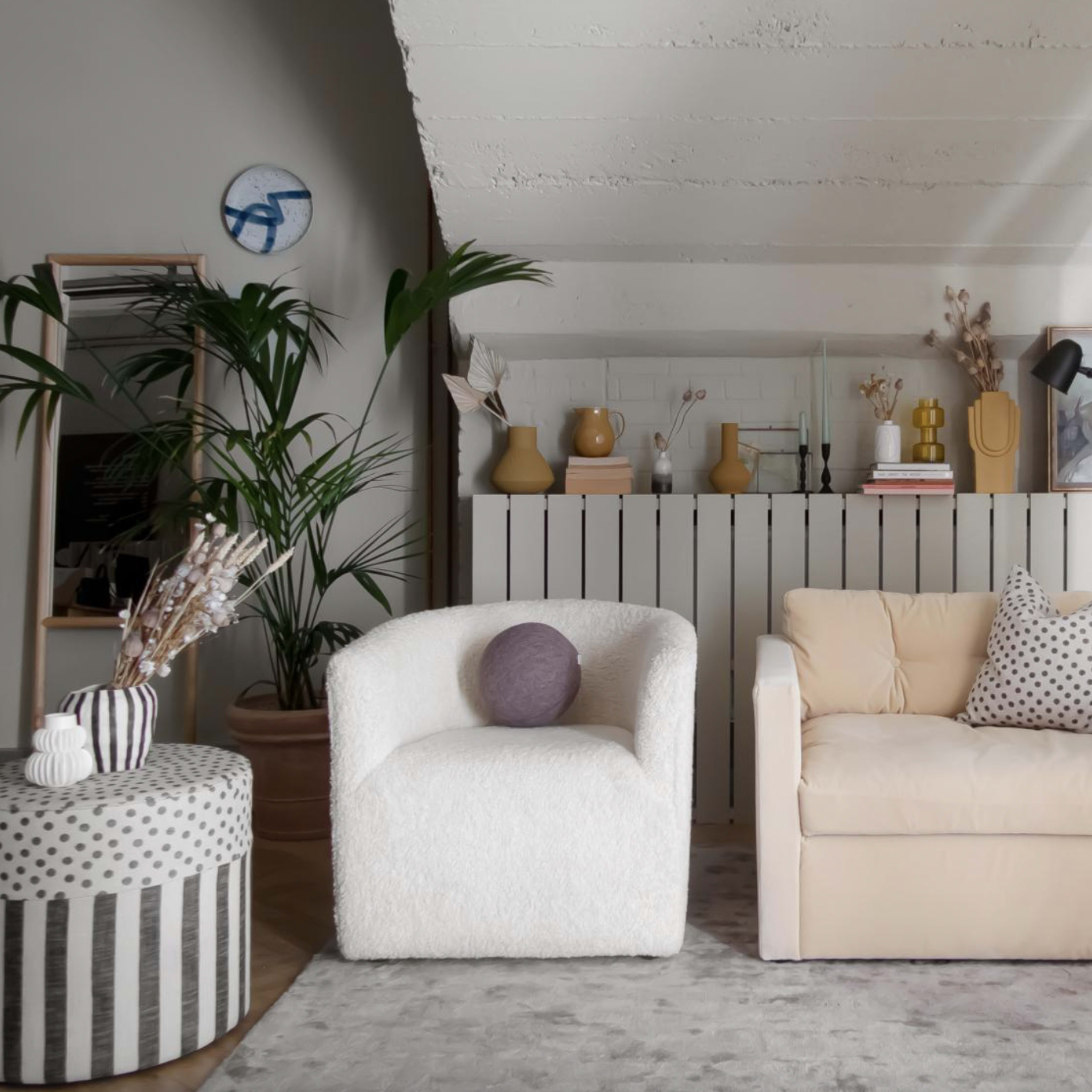 A cozy living room featuring two armchairs, a round striped table, and a light-colored sofa, with plants and decorative objects arranged on a low shelf.