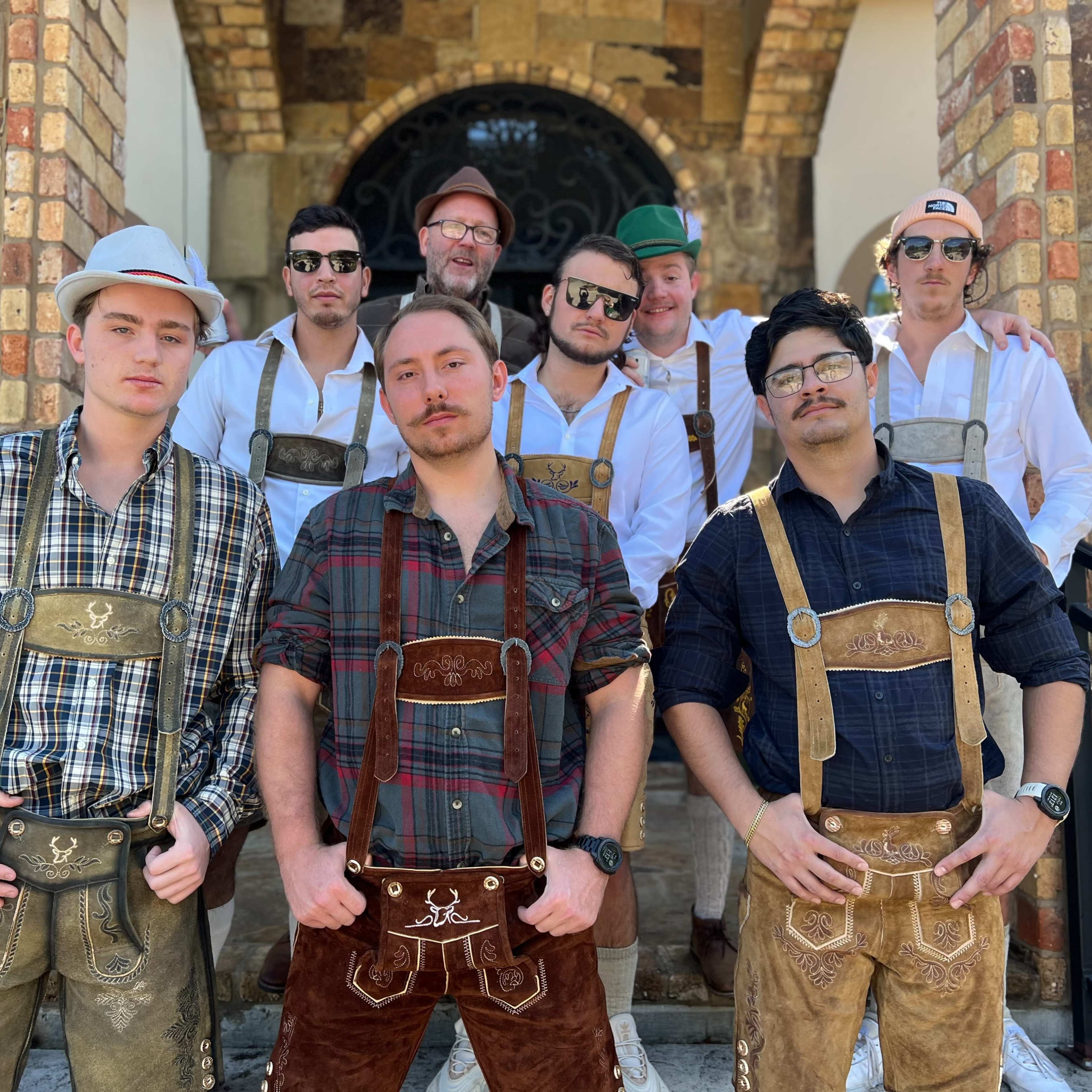 A group of nine men wearing traditional Bavarian attire, including lederhosen and hats, poses together on a staircase outside a stone building.