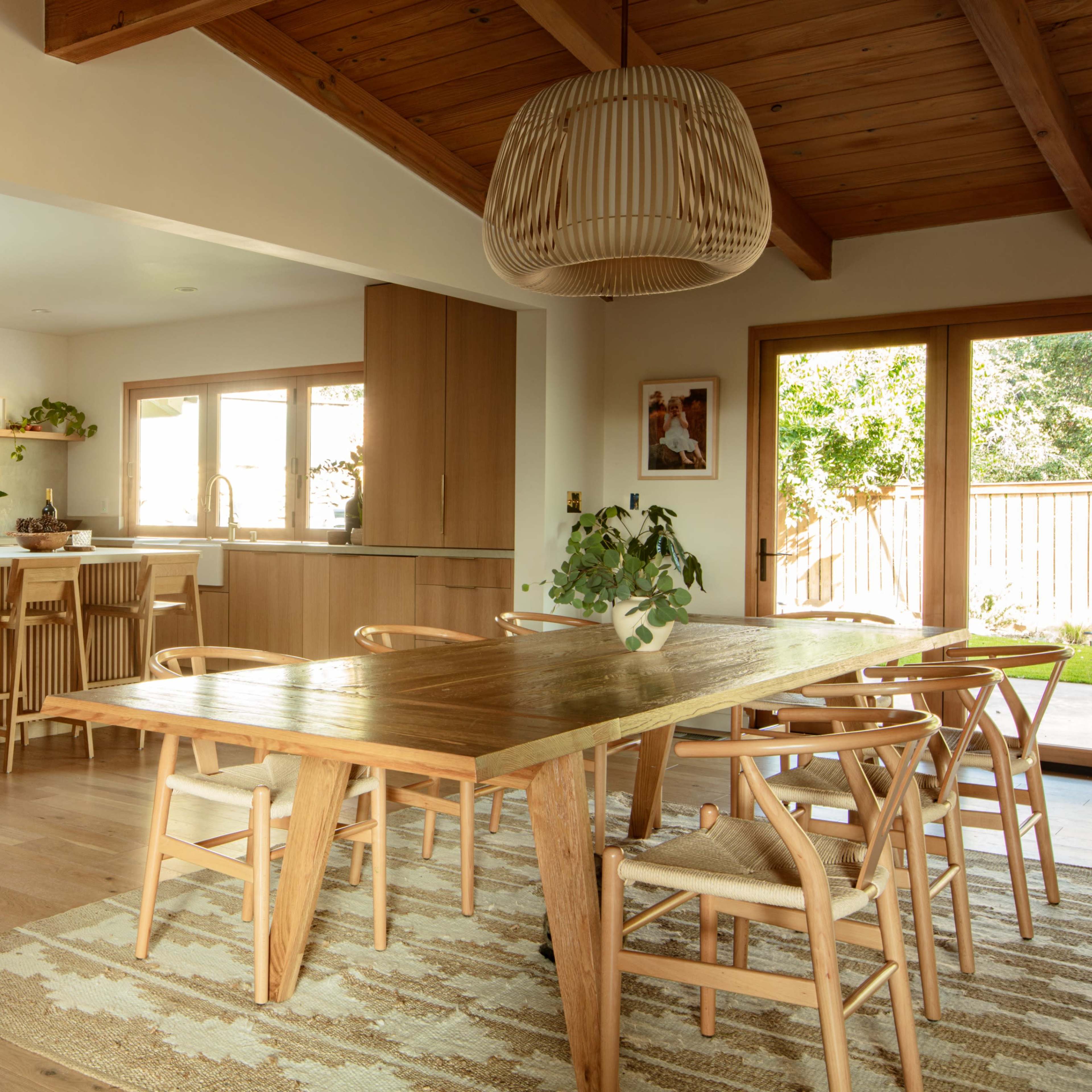 A modern kitchen and dining area with wooden finishes, a large dining table, and natural light coming through large windows.