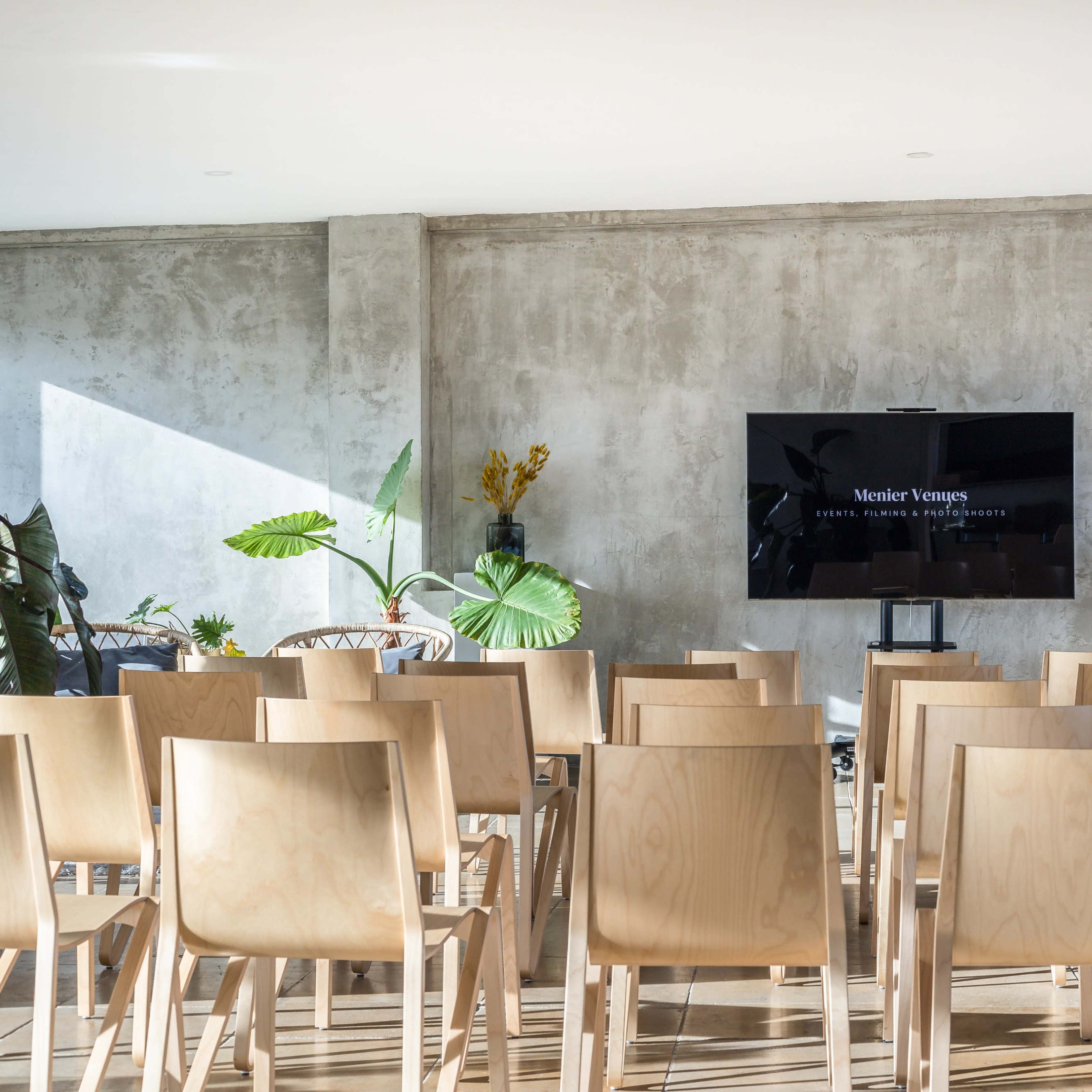 A rows of wooden chairs face a large television screen mounted on a concrete wall in a minimalistic conference room.