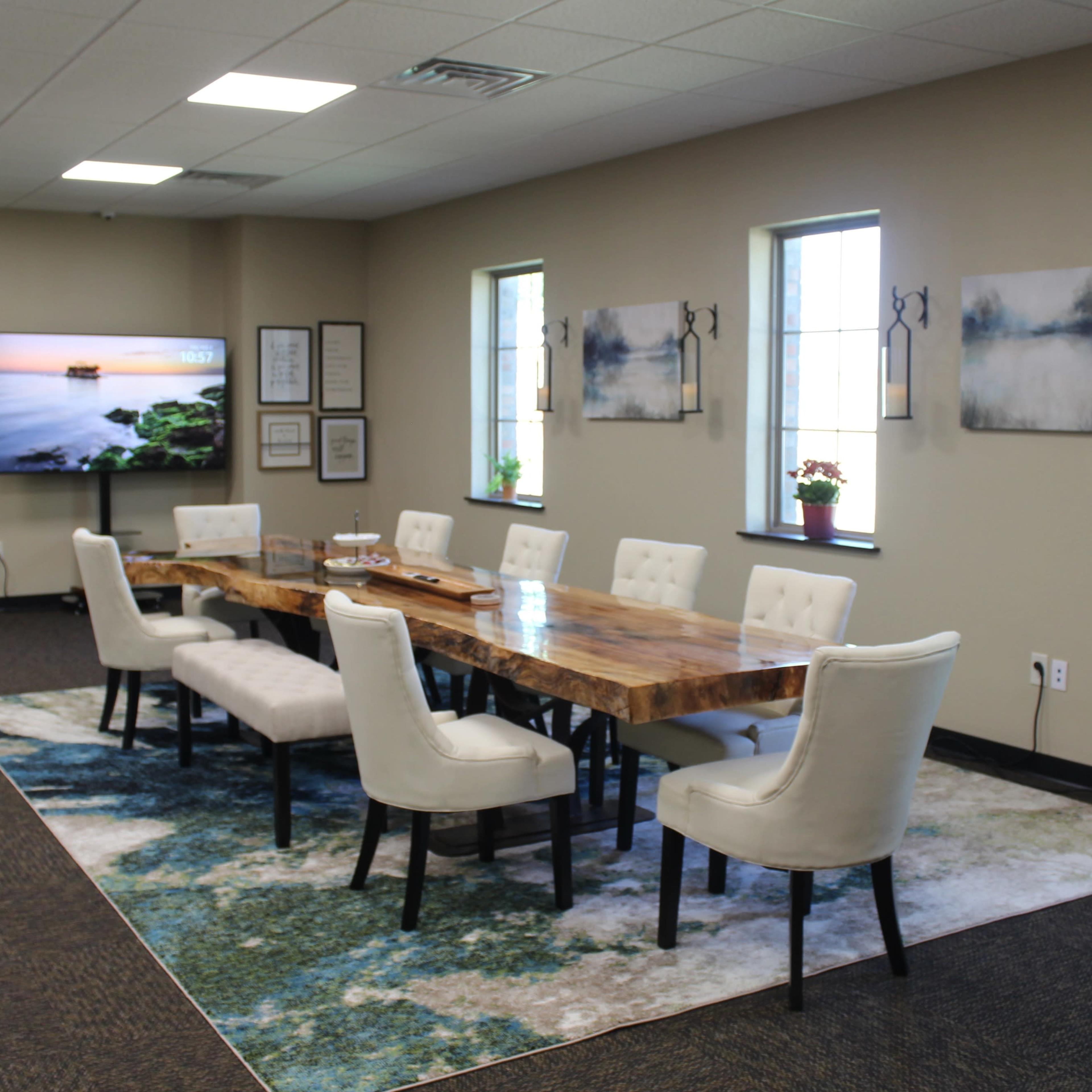 A long wooden table with white upholstered chairs is set in a well-lit meeting room, featuring windows and a large television screen on one wall.