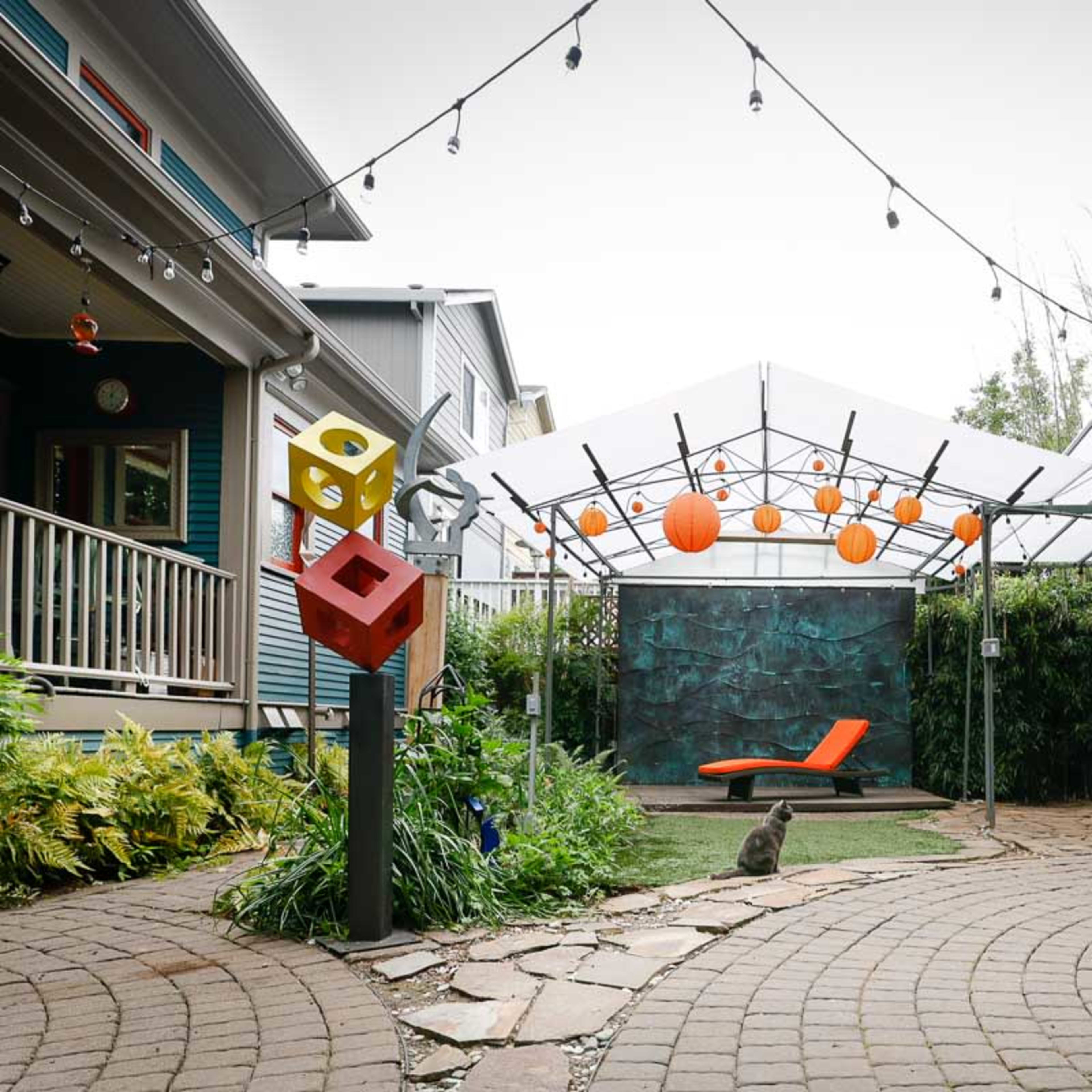 A landscaped outdoor space featuring a patio with a geometric sculpture, a red wall, an orange lounge chair, and string lights overhead.