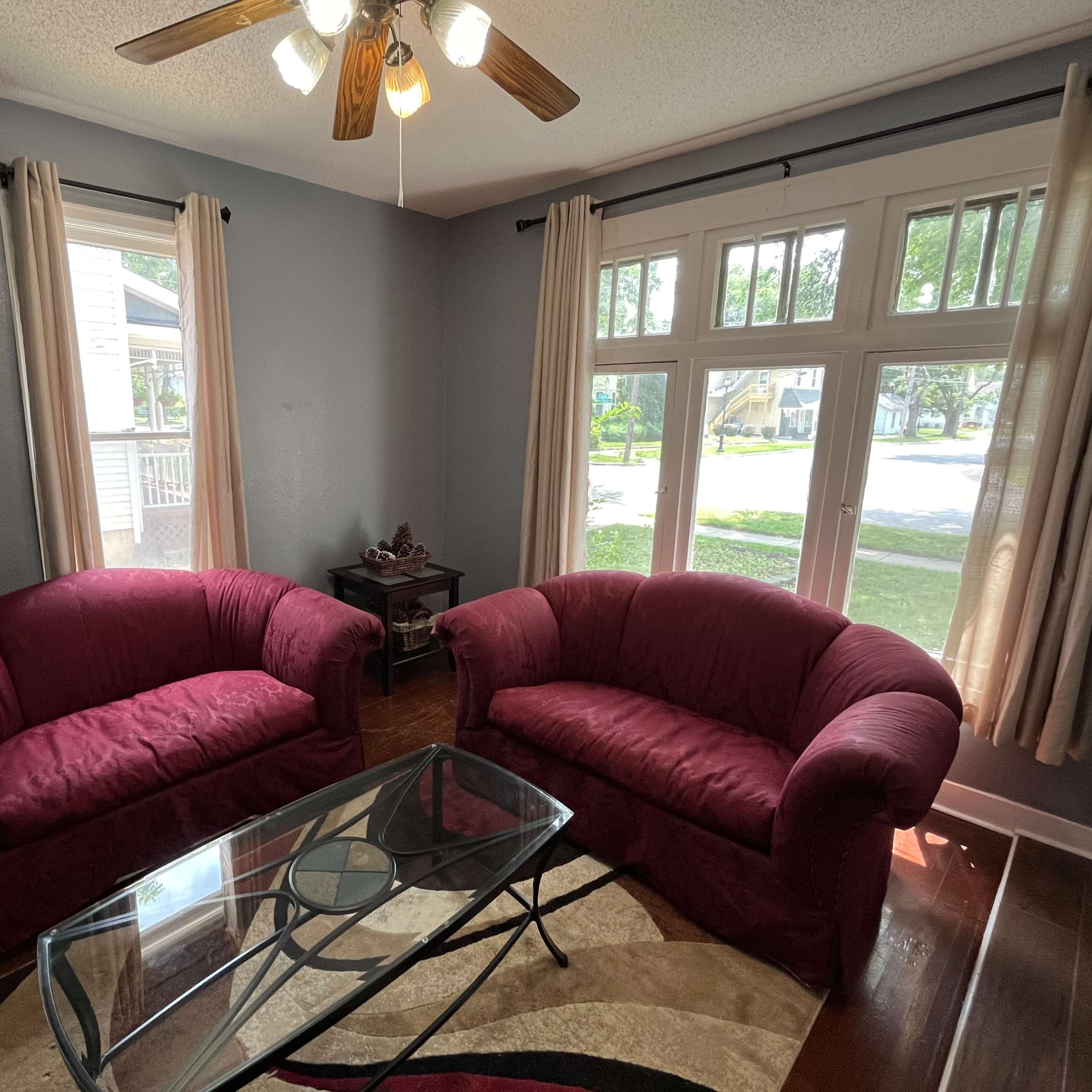 A living room with two red upholstered chairs, a glass coffee table, and large windows allowing natural light to enter.