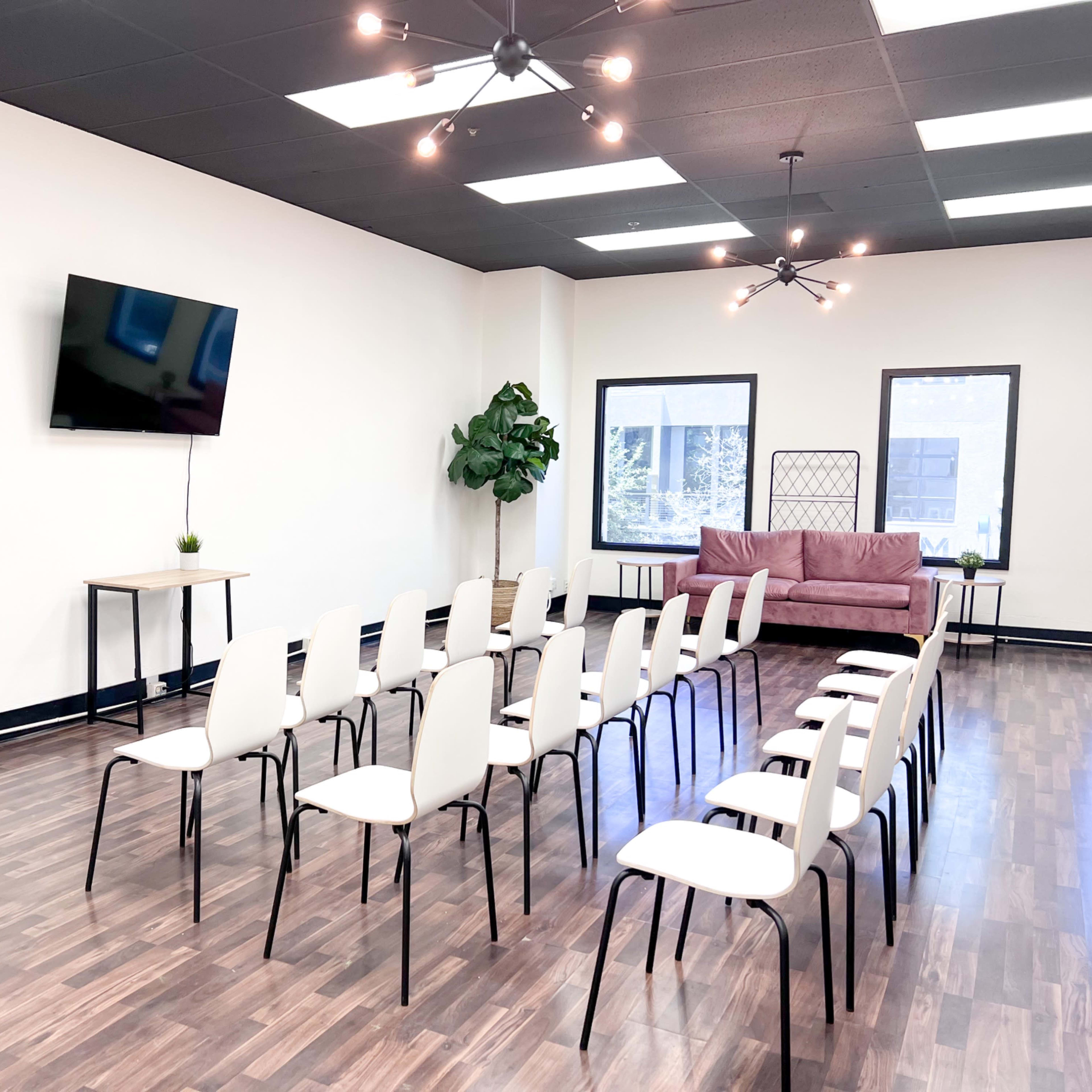 A conference room with a row of white chairs arranged facing a blank easel, a television mounted on the wall, and a couch in the background.