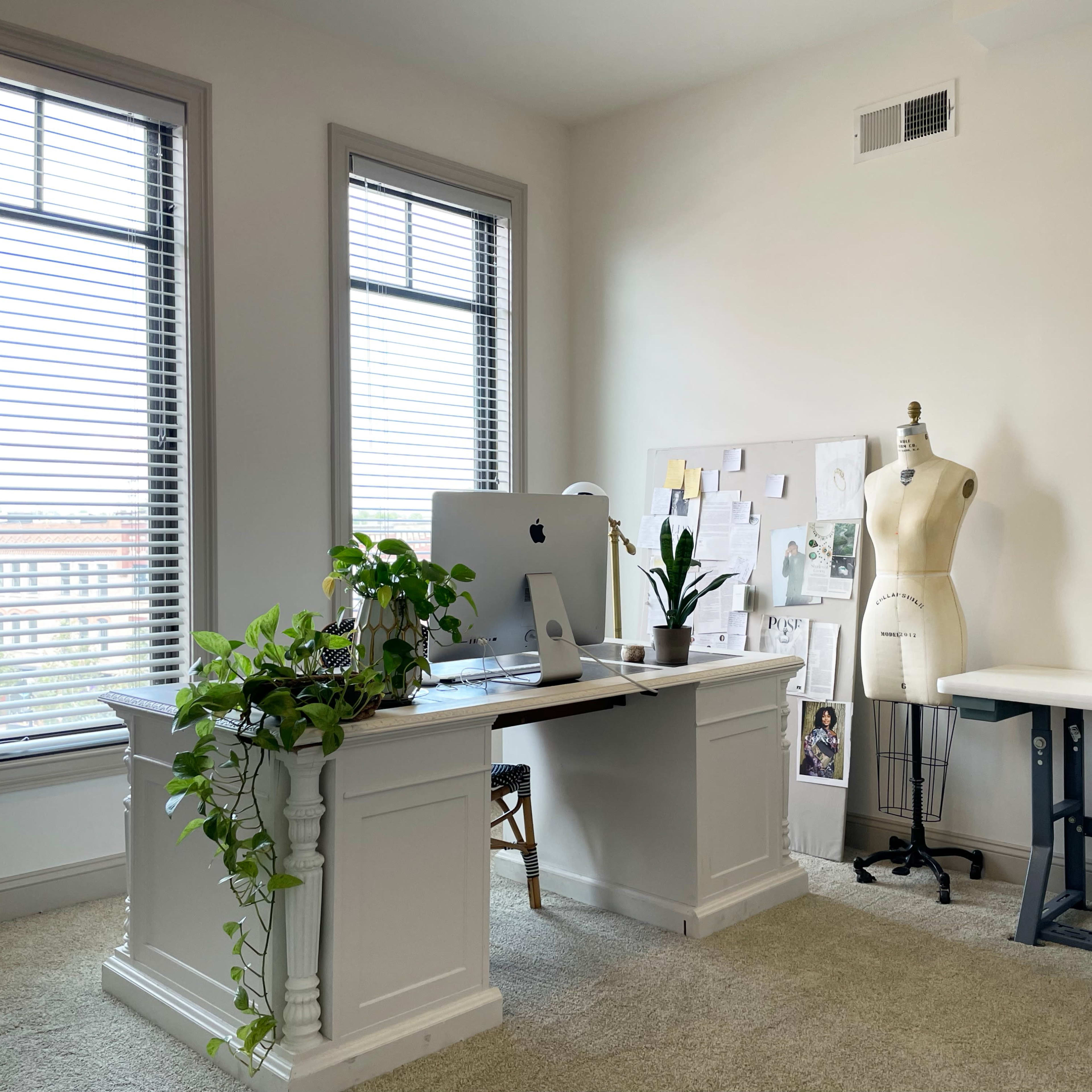 A bright office space features a desk with a computer, a sewing machine, and a dress form, alongside large windows and a plant on the desk.