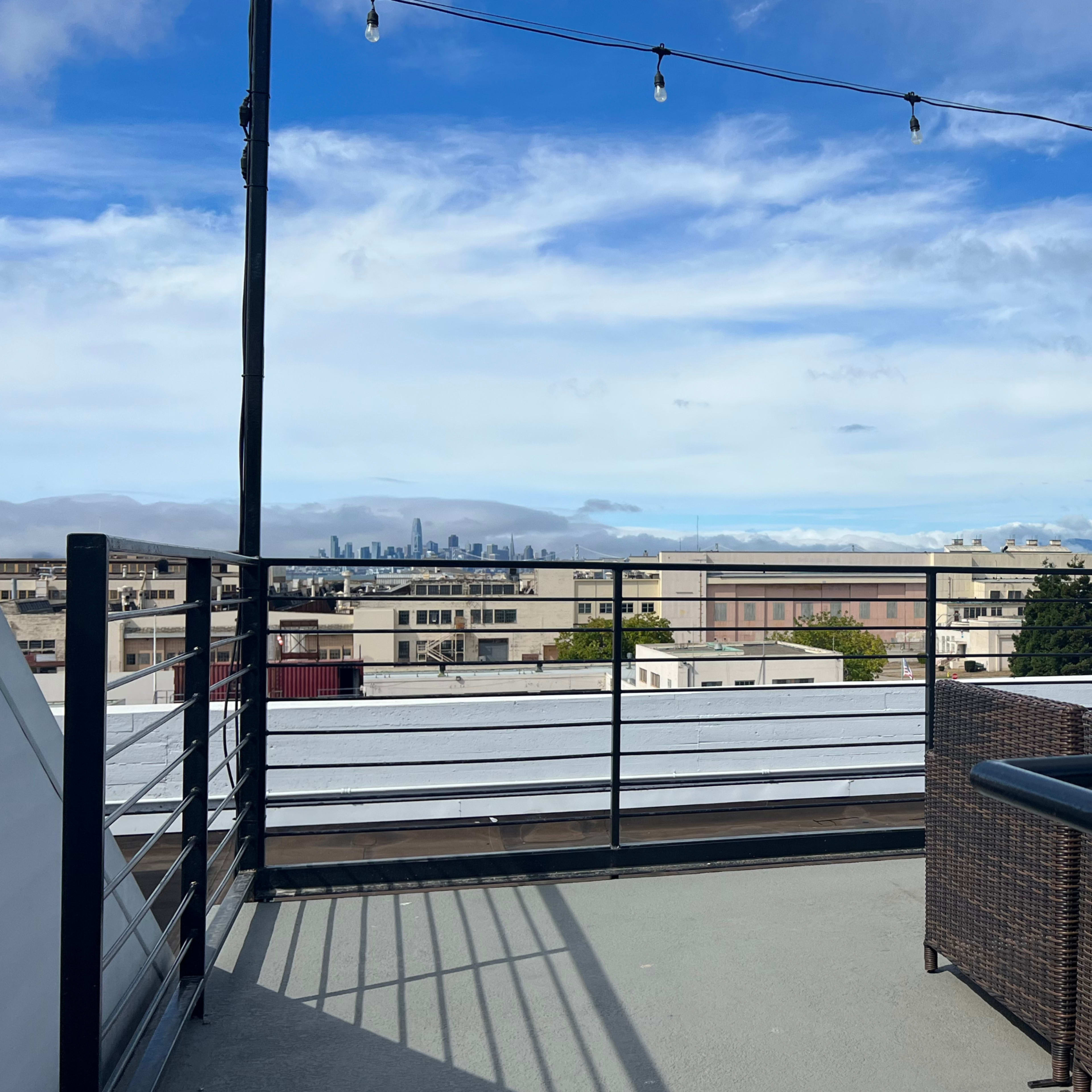 A rooftop deck with string lights, overlooking city buildings and a cloudy sky.