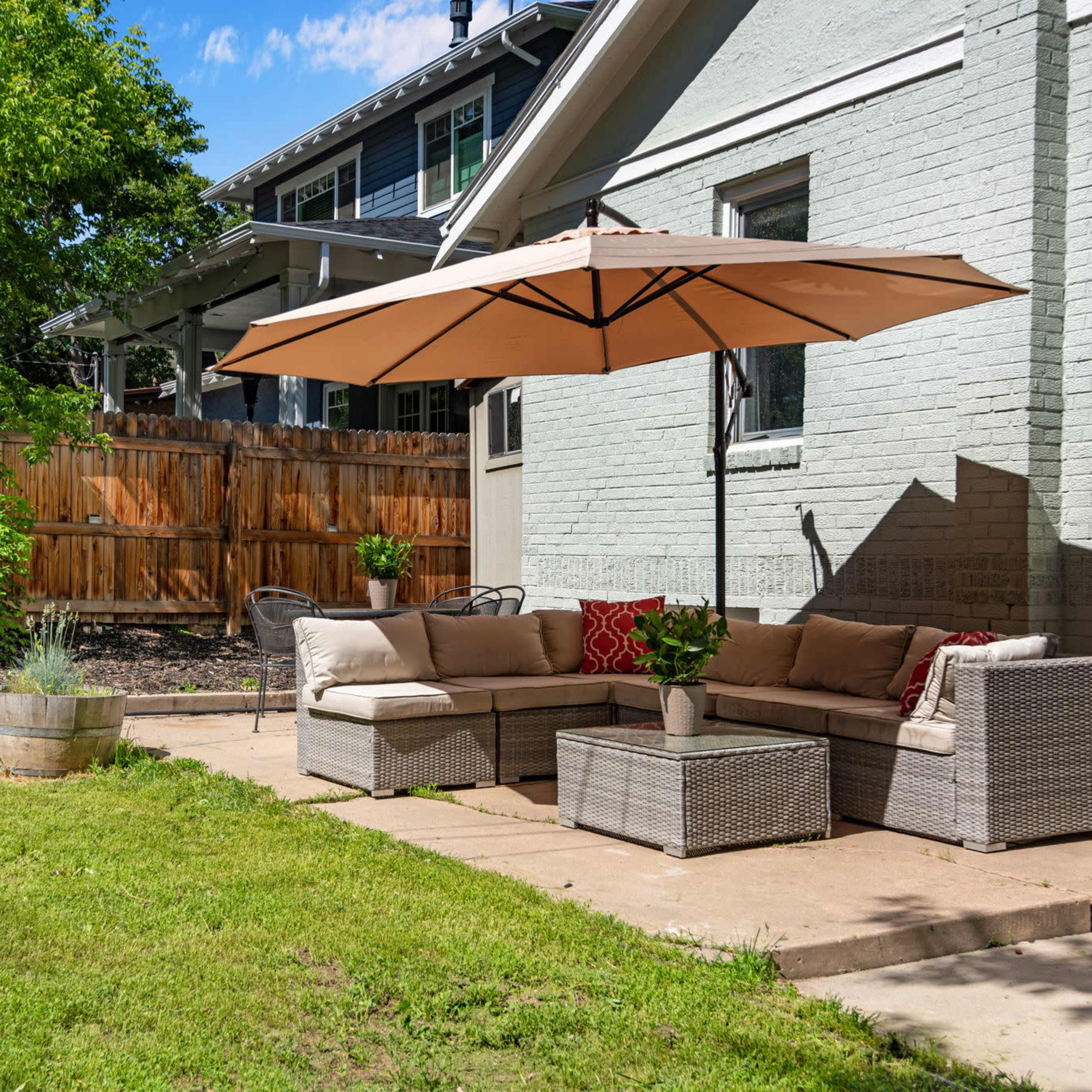 A patio area with a sectional sofa under an umbrella, a barbecue grill nearby, and a green lawn surrounded by a wooden fence.