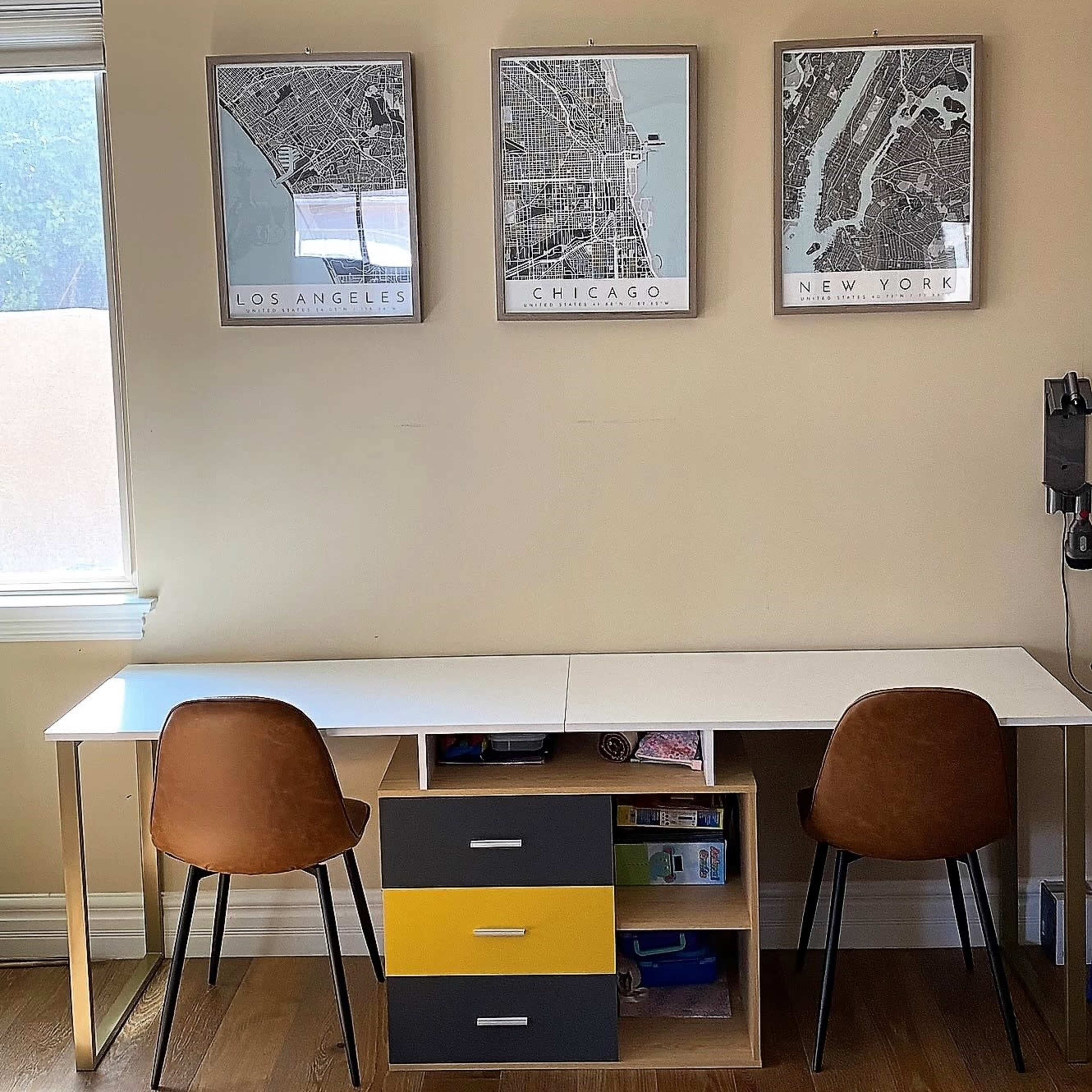 A minimalist workspace with a long white desk, two brown chairs, and a storage unit with yellow, gray, and white drawers, framed city map posters of Los Angeles, Chicago, and New York hanging on the wall.