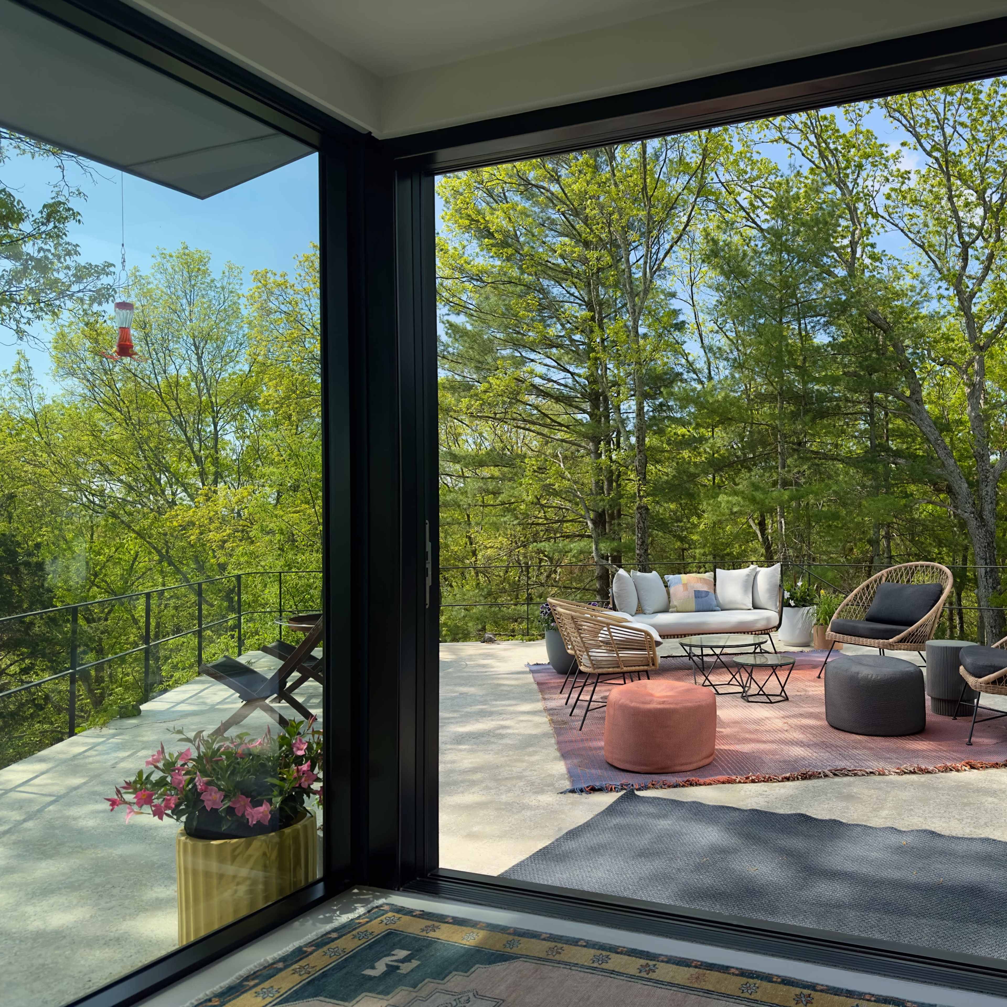 A modern outdoor seating area with wicker chairs and a circular table, surrounded by trees and greenery, viewed through large glass windows.