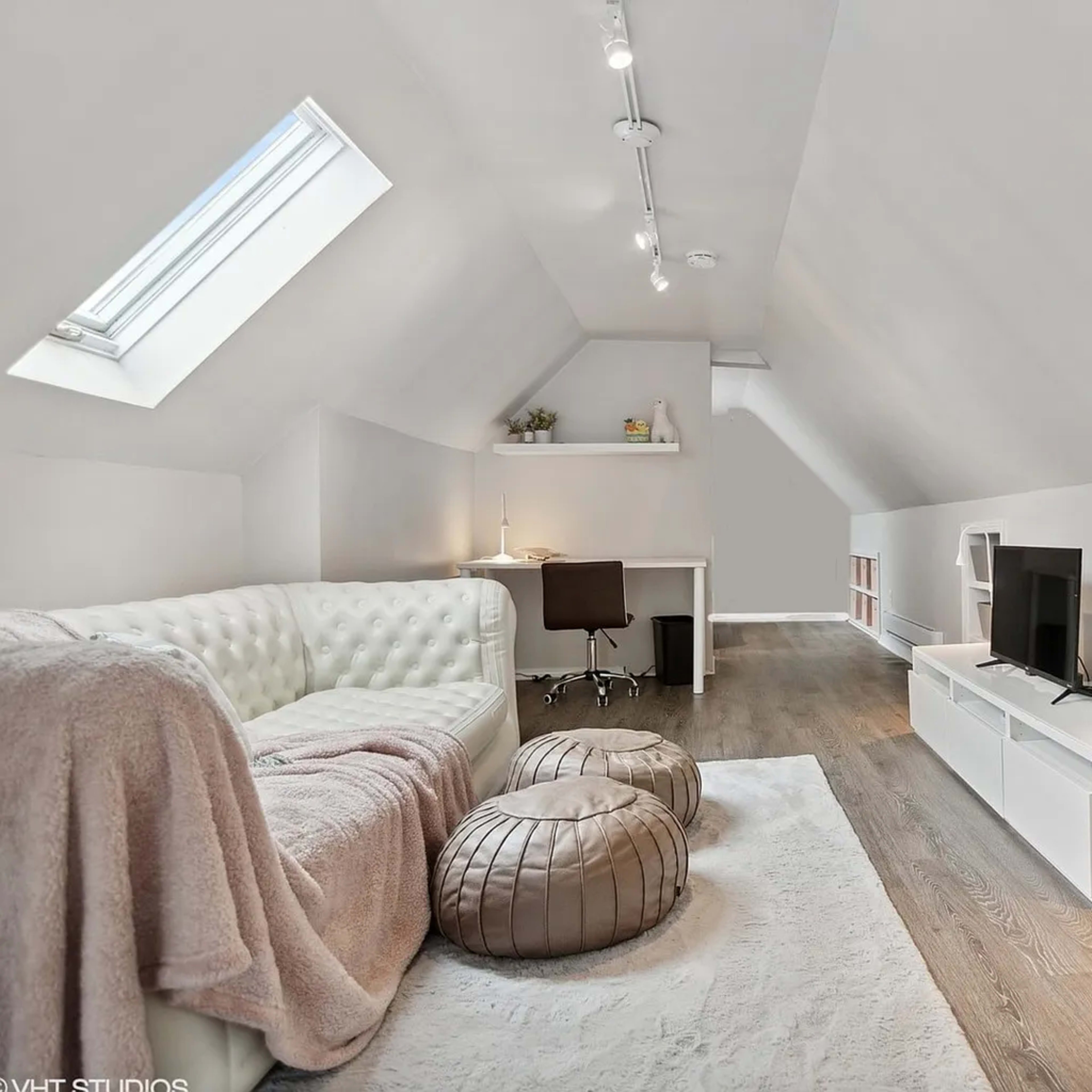 A modern attic space with a white sofa, a desk with a chair, a TV stand, and two large cushioned poufs.