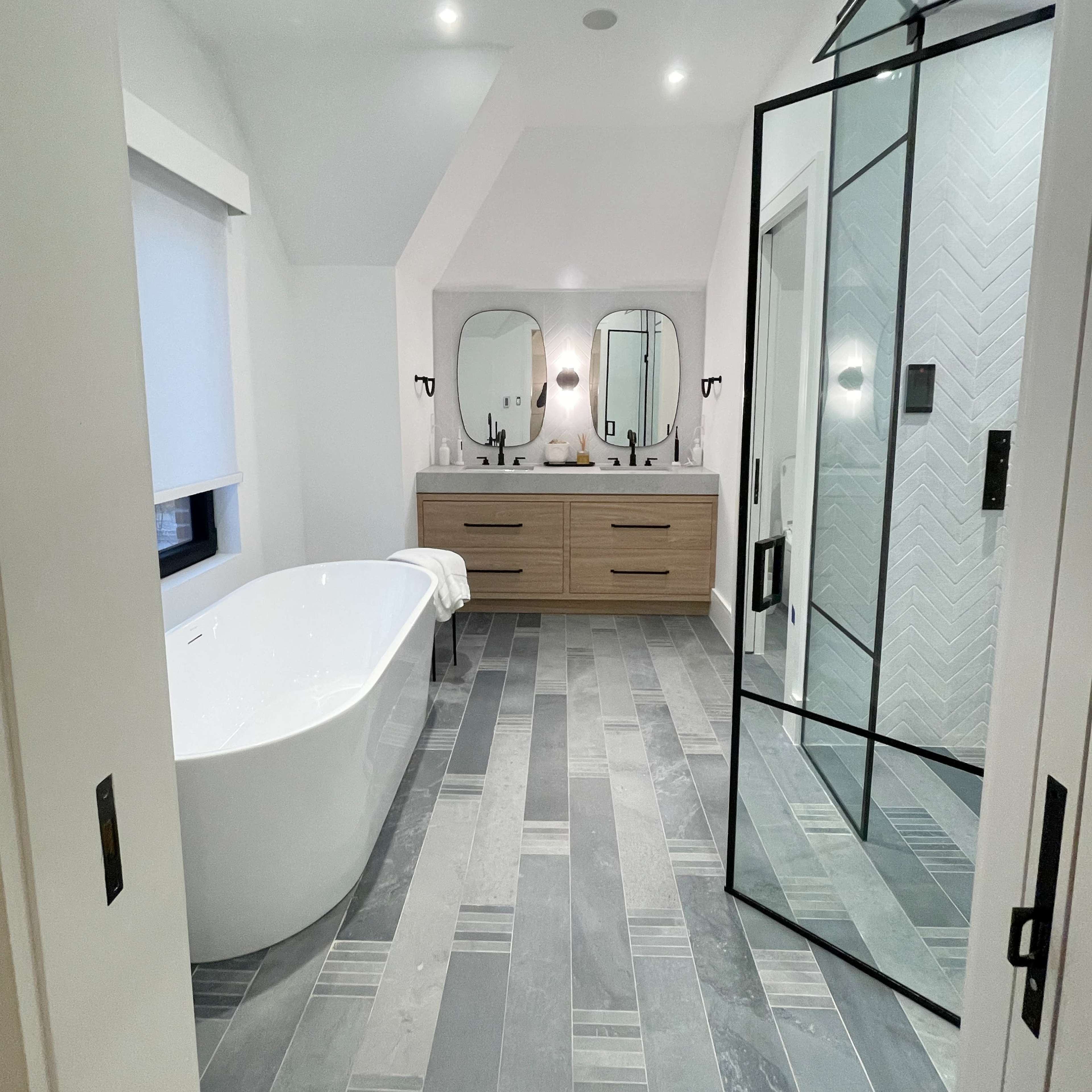 A modern bathroom features a freestanding bathtub, dual sinks with mirrors, and a glass shower enclosure on tiled flooring.