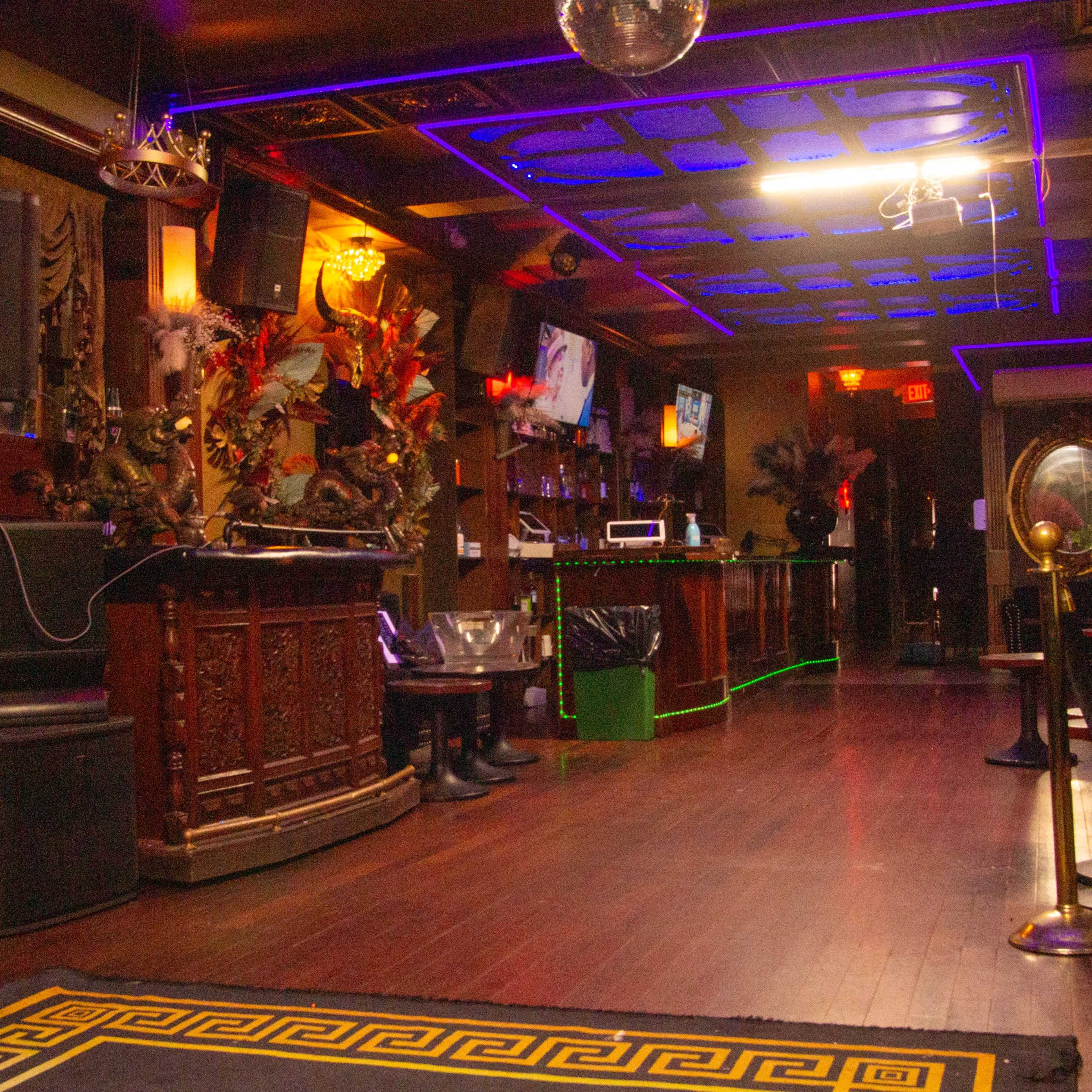 A dimly lit bar with ornate decor, a wooden bar counter, and neon blue lighting illuminating the ceiling and walls.