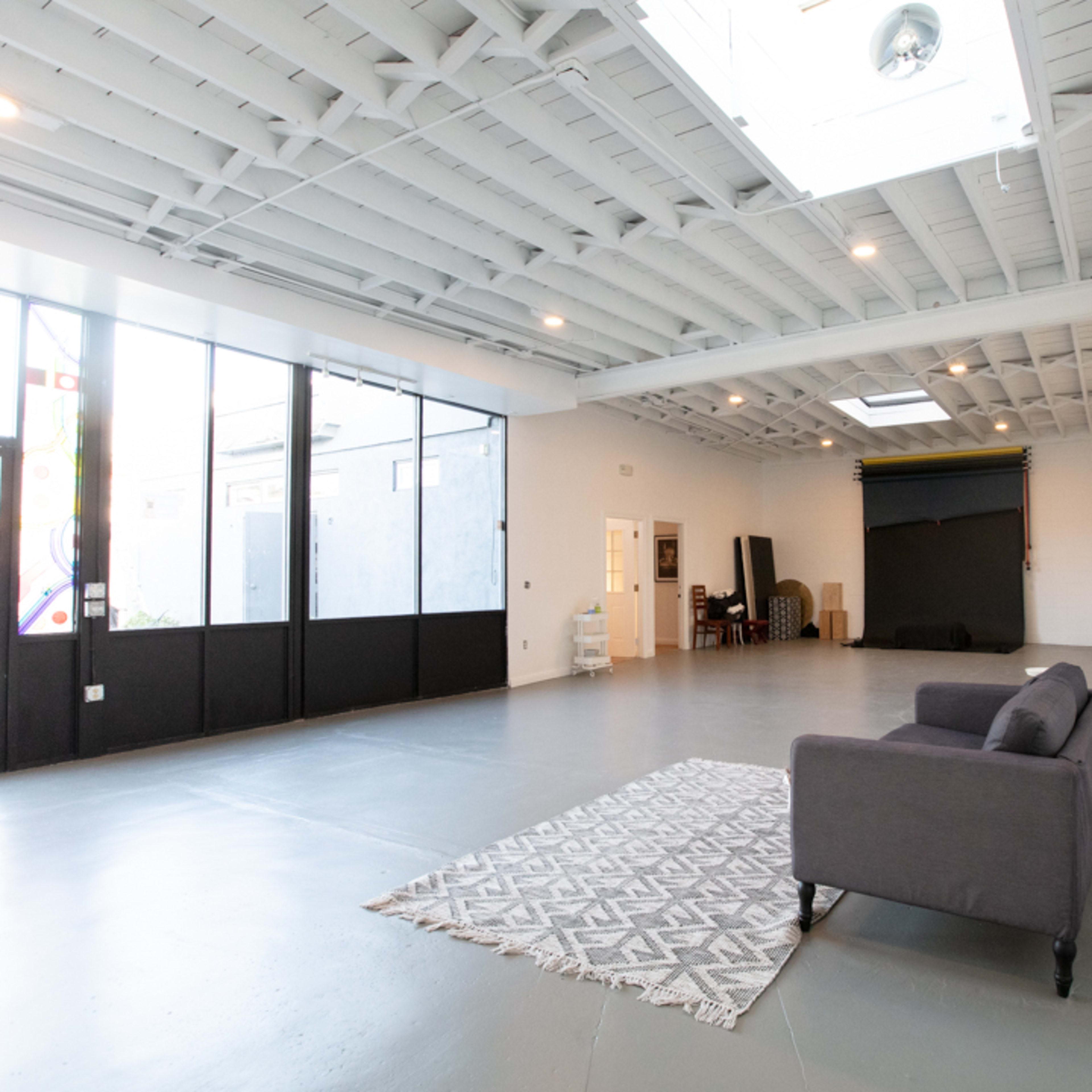 A spacious, empty studio with large windows, a gray couch, and a patterned rug on a concrete floor.