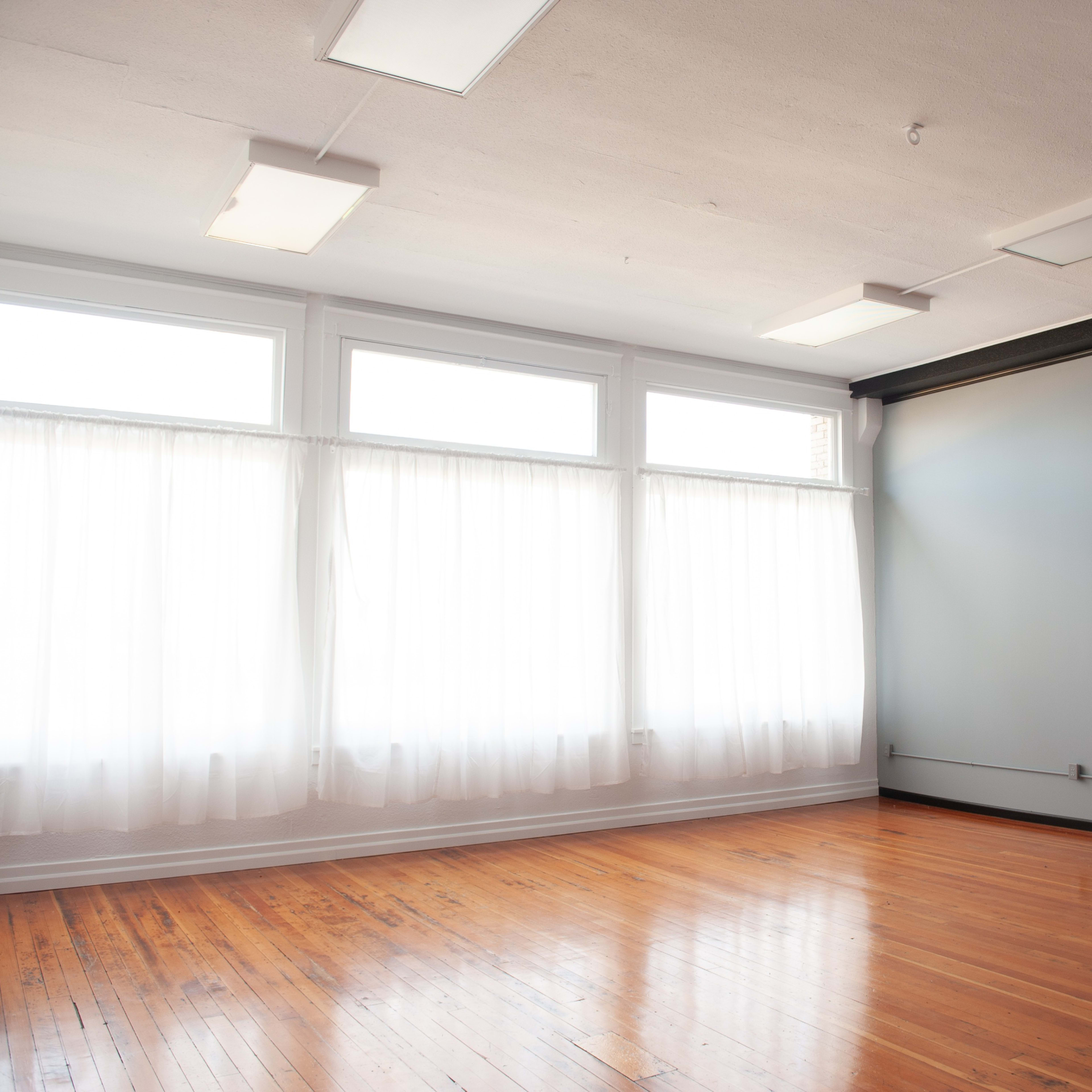 An empty room with large windows covered by sheer curtains and polished wooden floors.