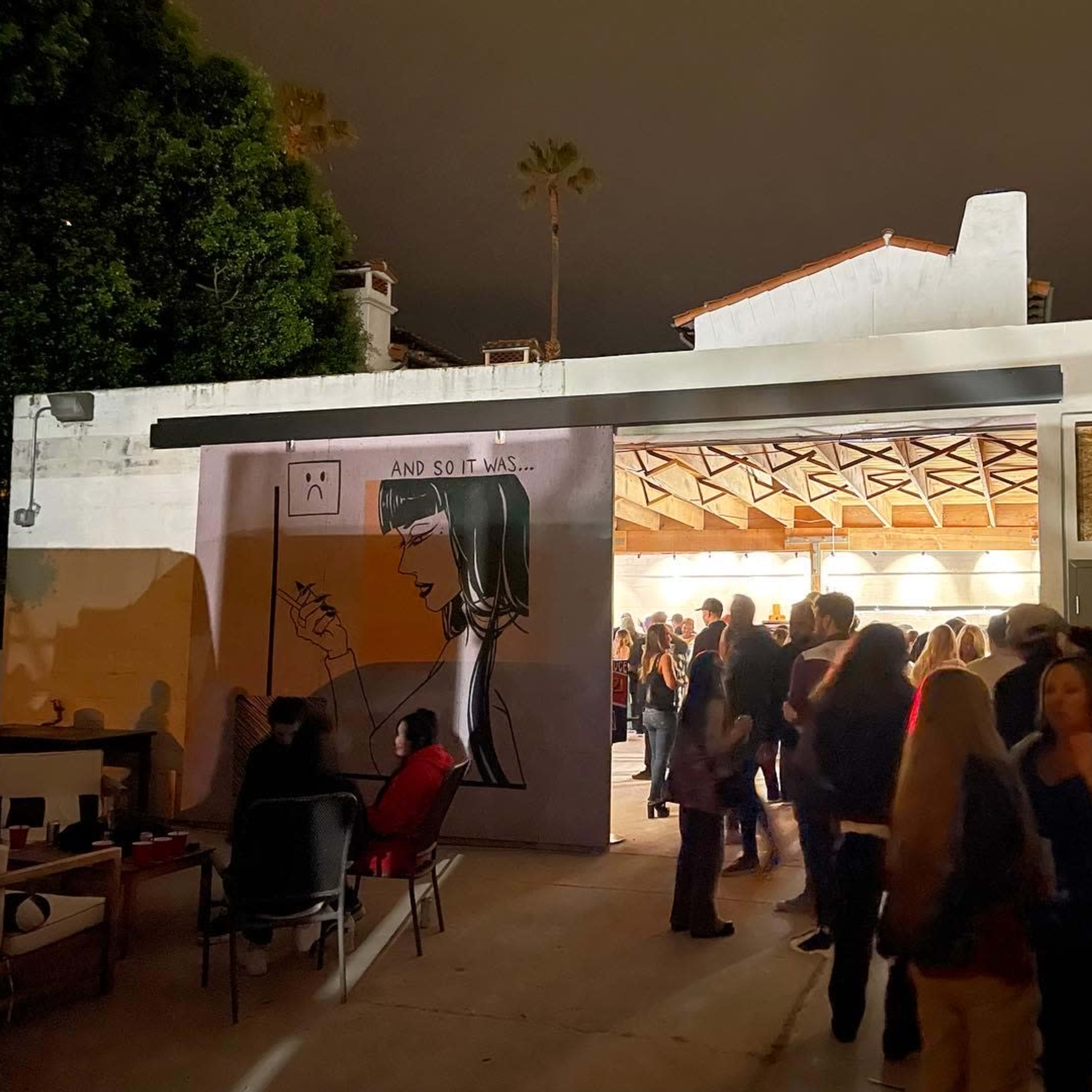 A crowd gathers outside a building featuring a large mural of a woman holding a pencil, under a night sky.