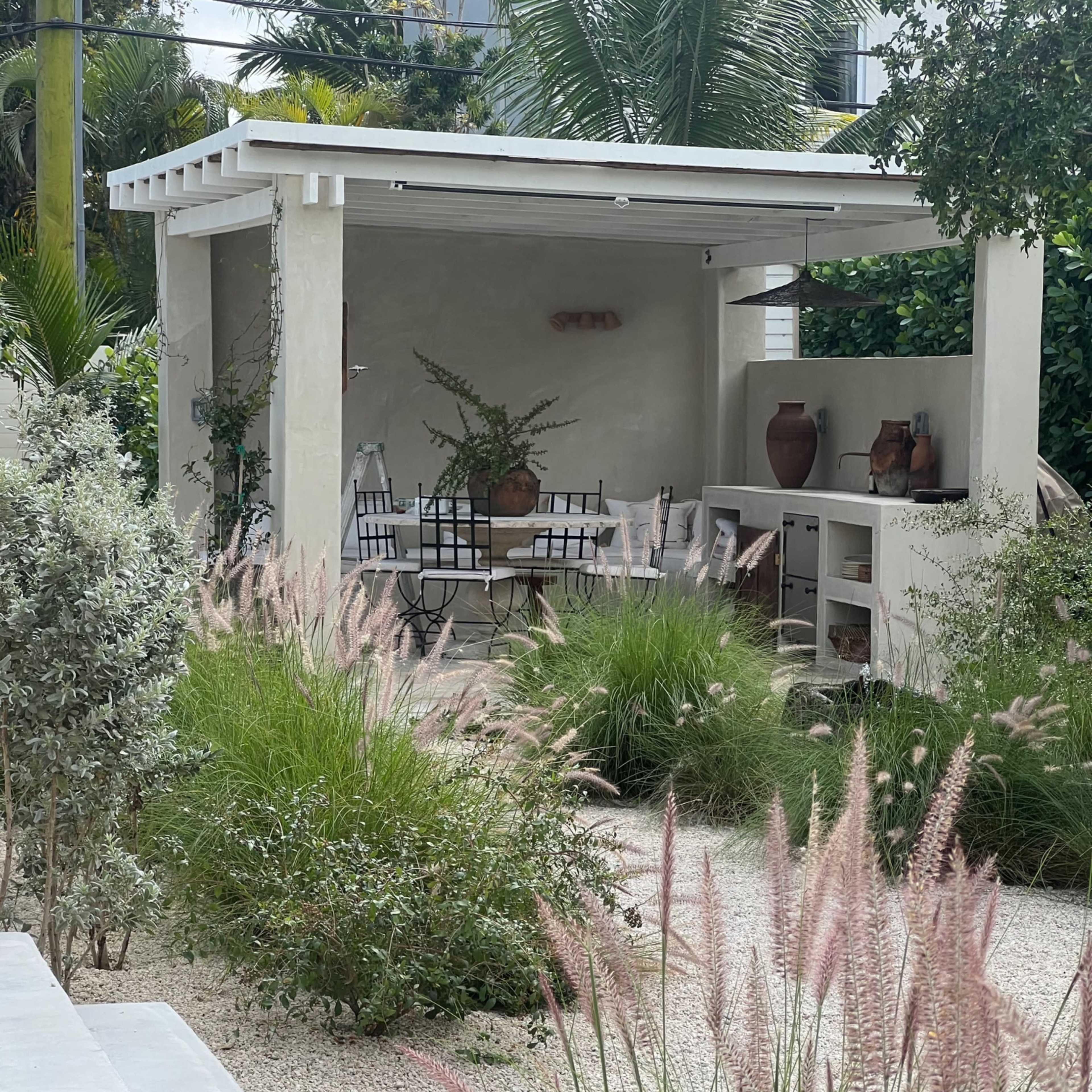 A modern outdoor pavilion with a dining area surrounded by lush greenery and ornamental grasses.