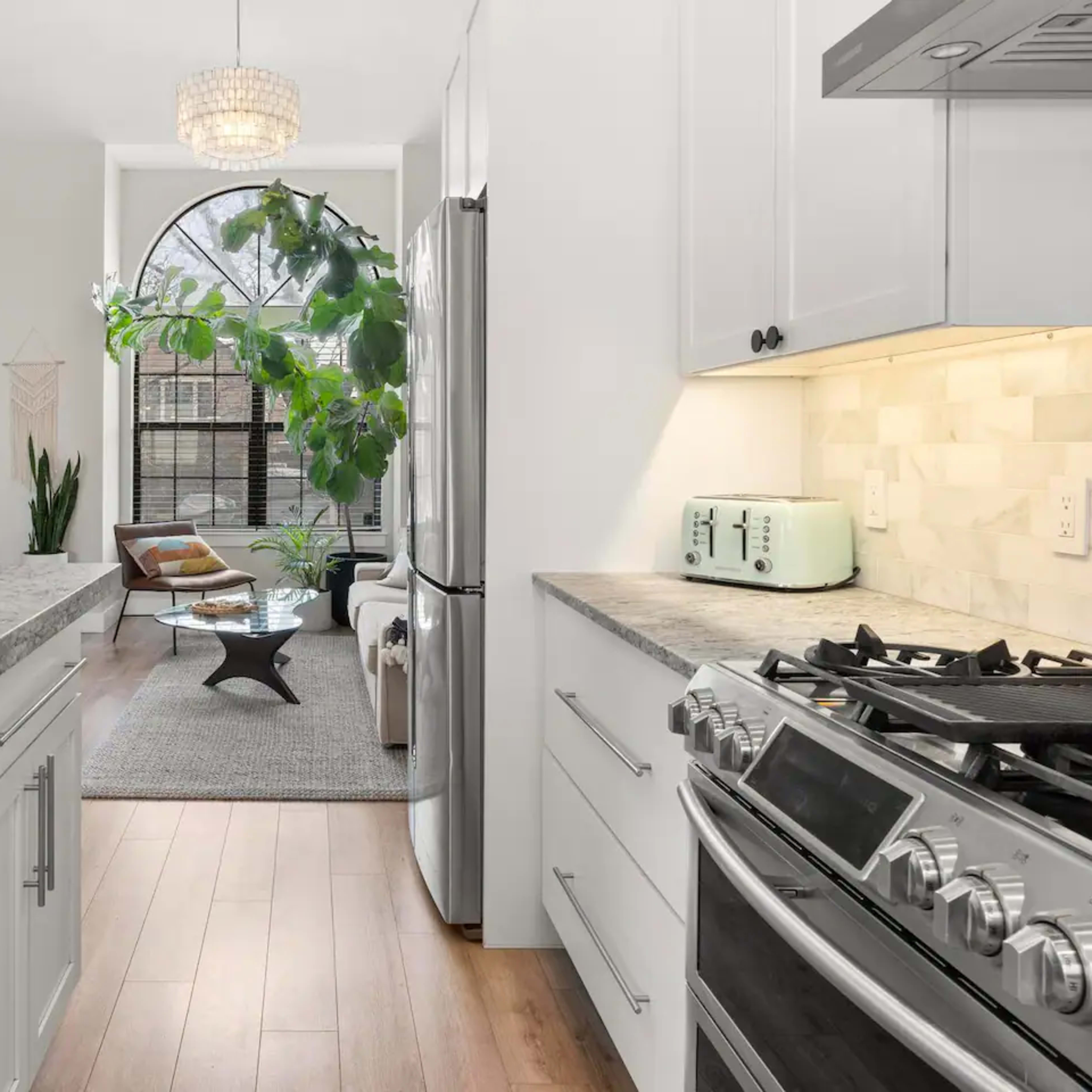 A modern kitchen features light-colored cabinets, a granite countertop, and a stainless steel gas stove, leading into a living area with a large window and indoor plants.