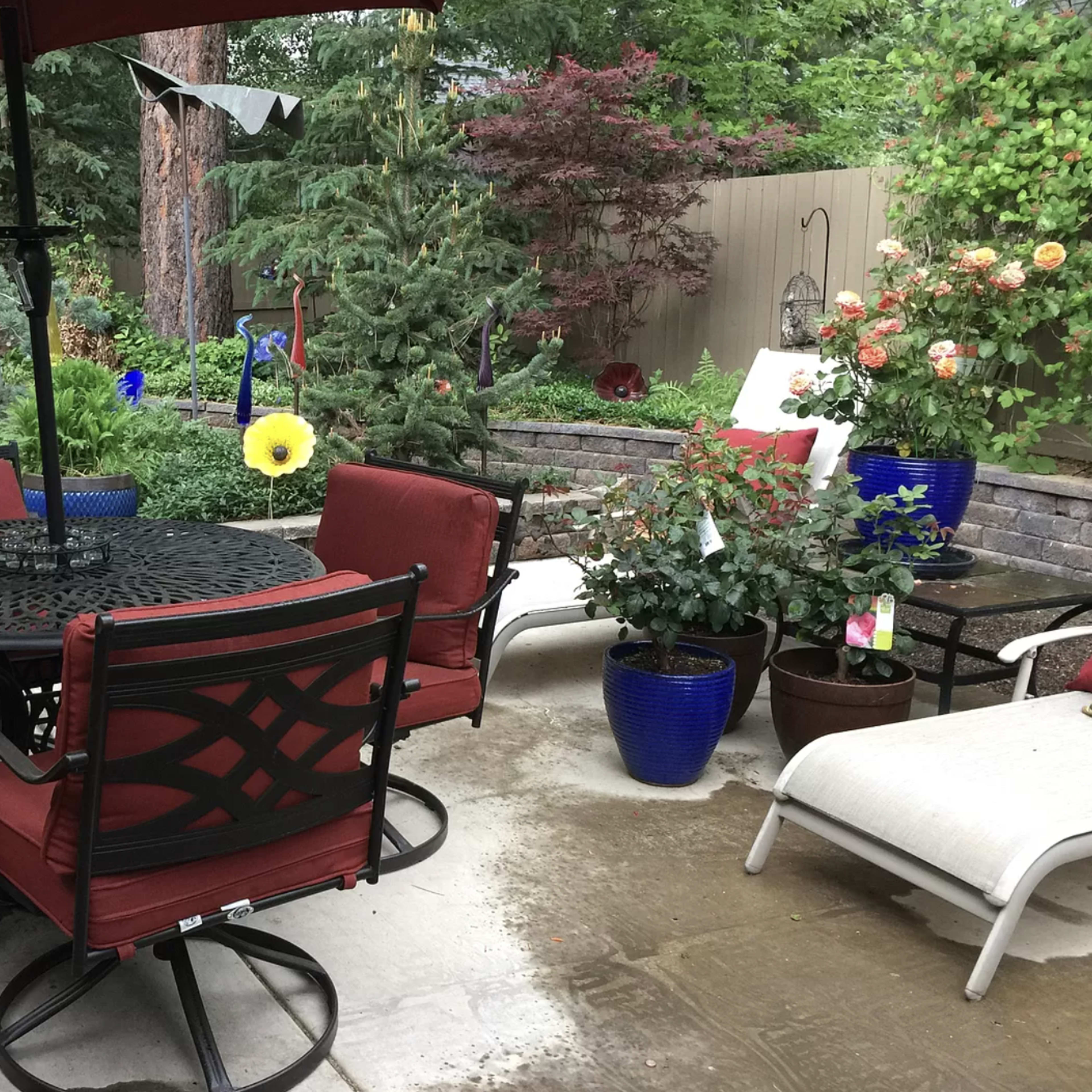 A patio with a round table and red chairs, surrounded by potted plants and a grill in the background.