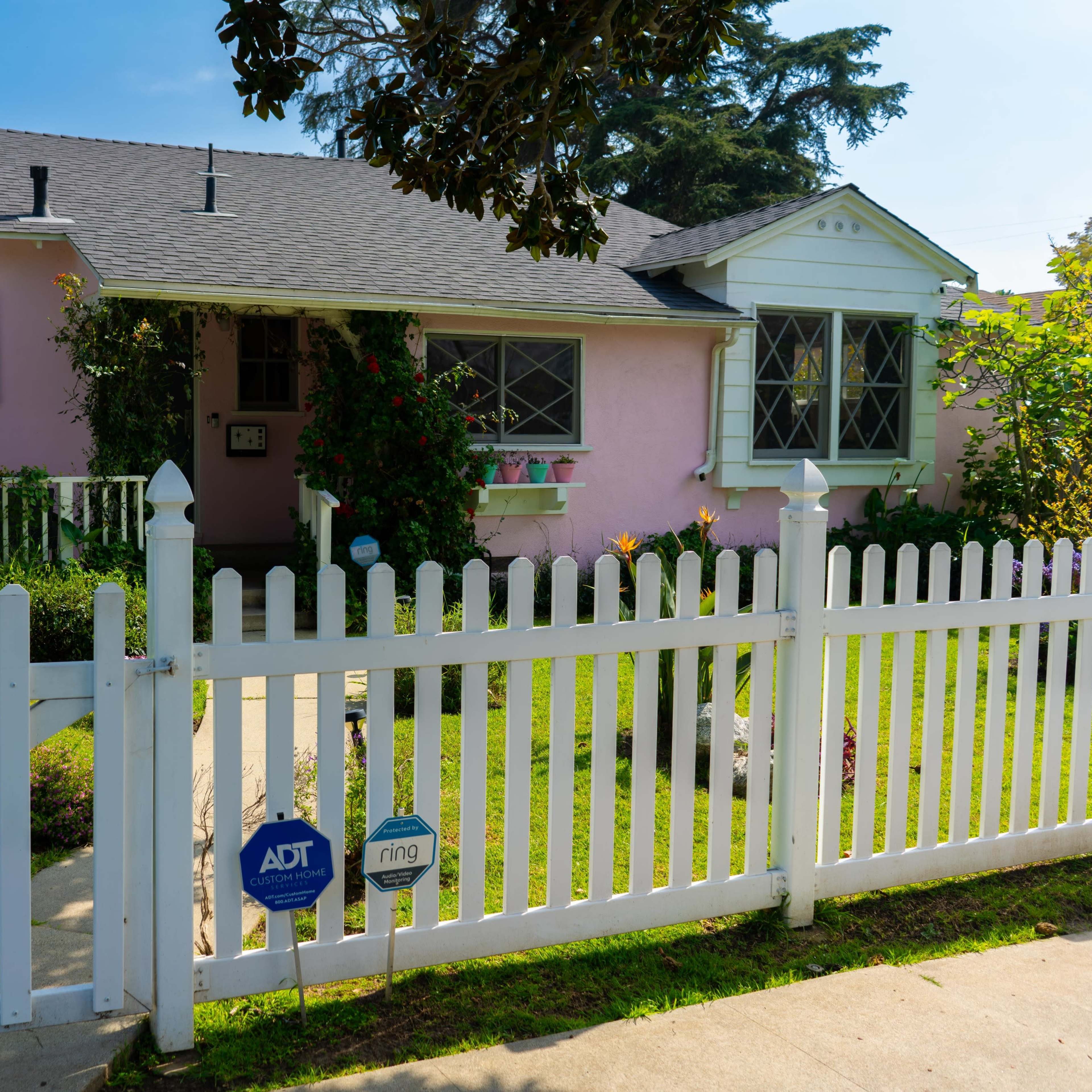 A pink house with white trim and a picket fence is surrounded by greenery and features a well-maintained front yard.