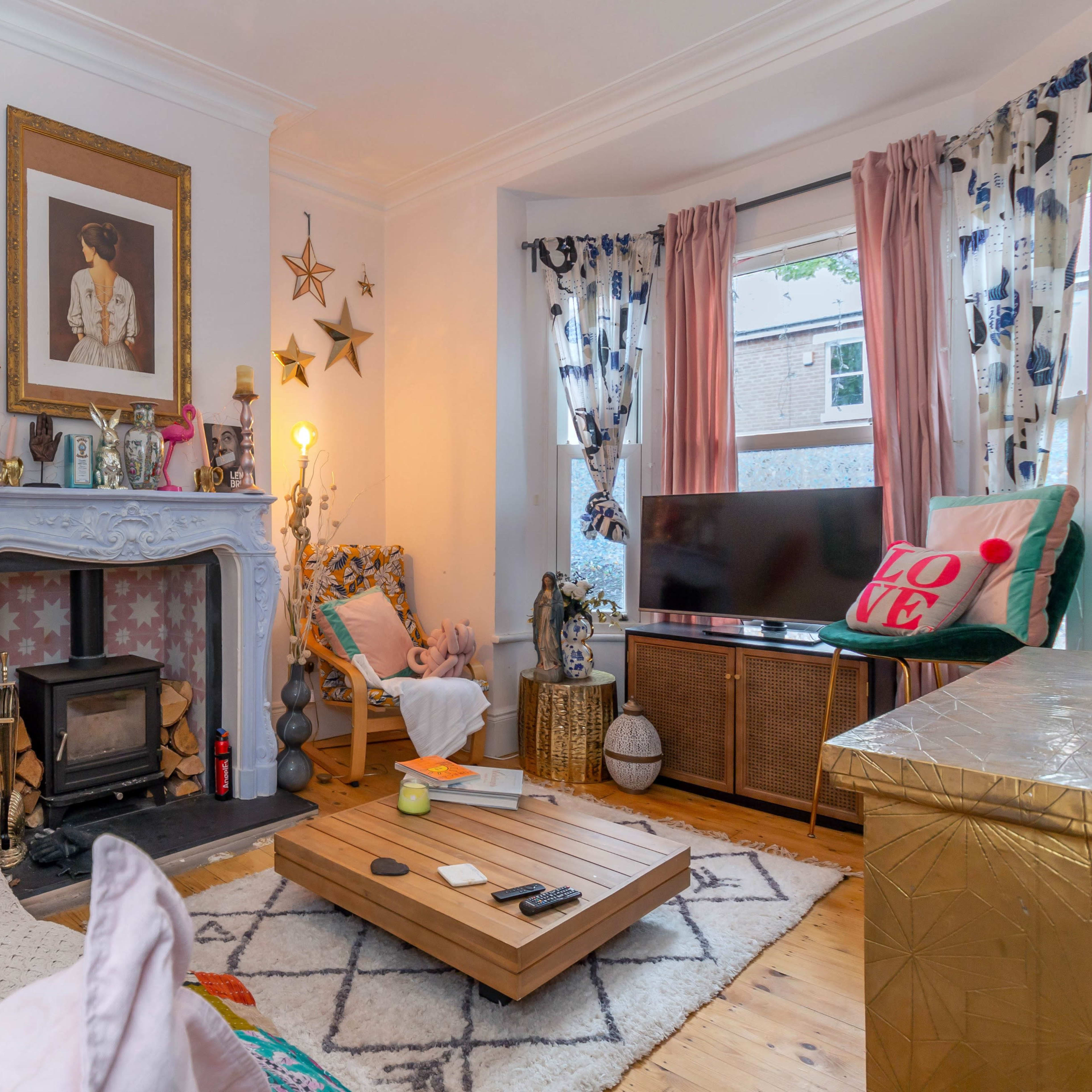A cozy living room featuring a pastel-colored sofa with decorative pillows, a wooden coffee table, a fireplace with a blue mantel, and large windows adorned with patterned curtains.