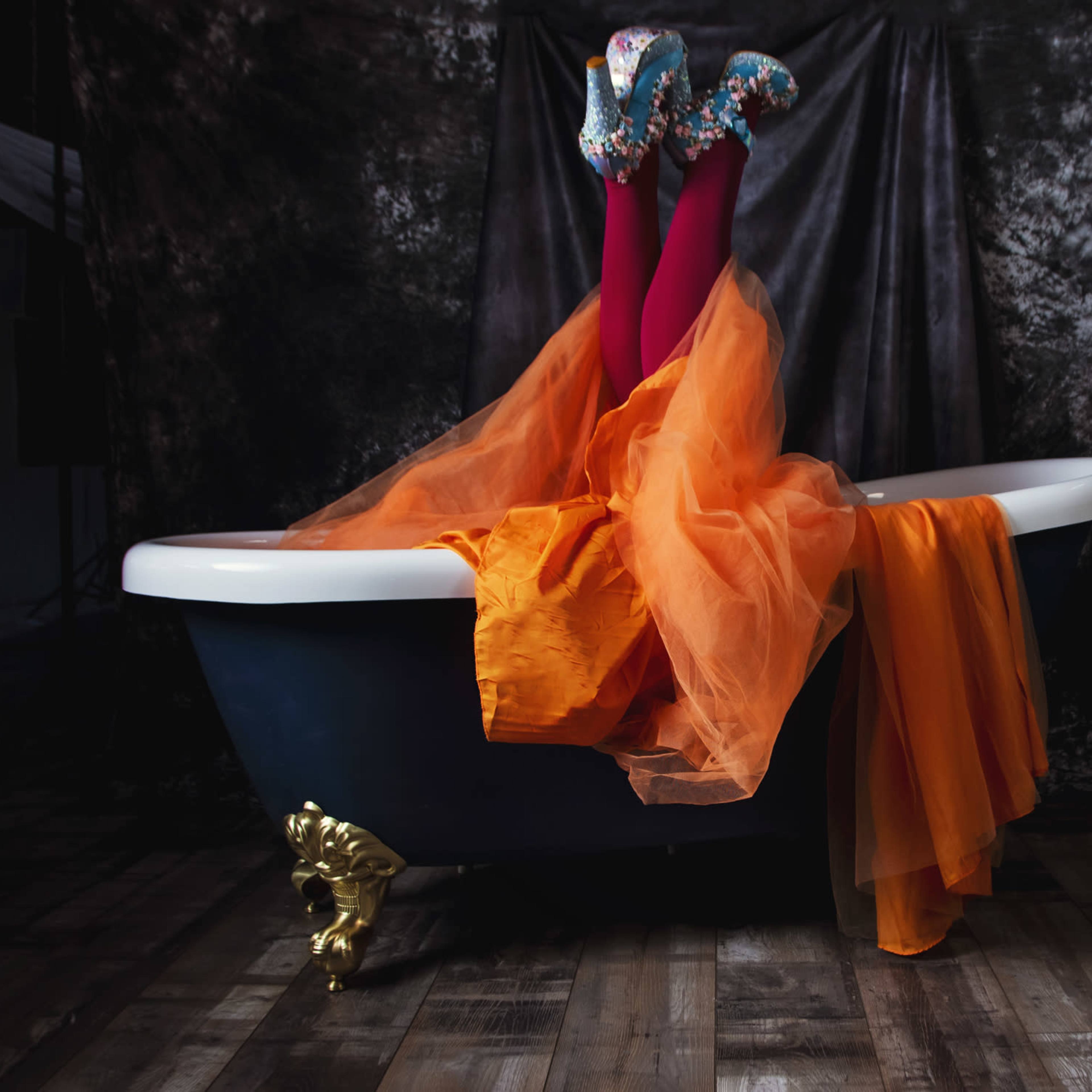 A pair of brightly clad legs with patterned shoes protrude from a vintage bathtub draped in orange fabric, set against a dark backdrop.