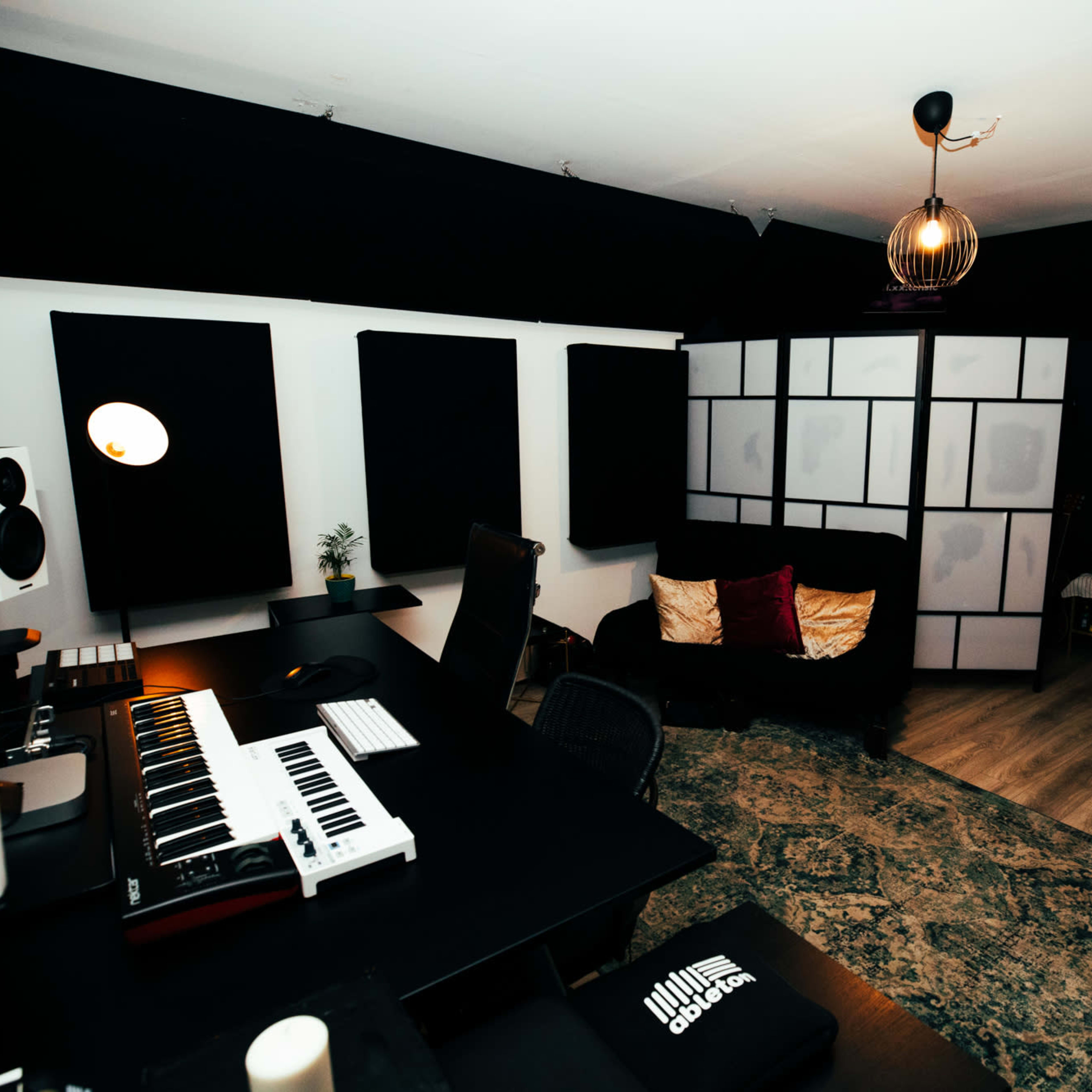 A modern music production studio featuring a keyboard on a desk, soundproof panels on the walls, and a seating area with a mirror and furniture.