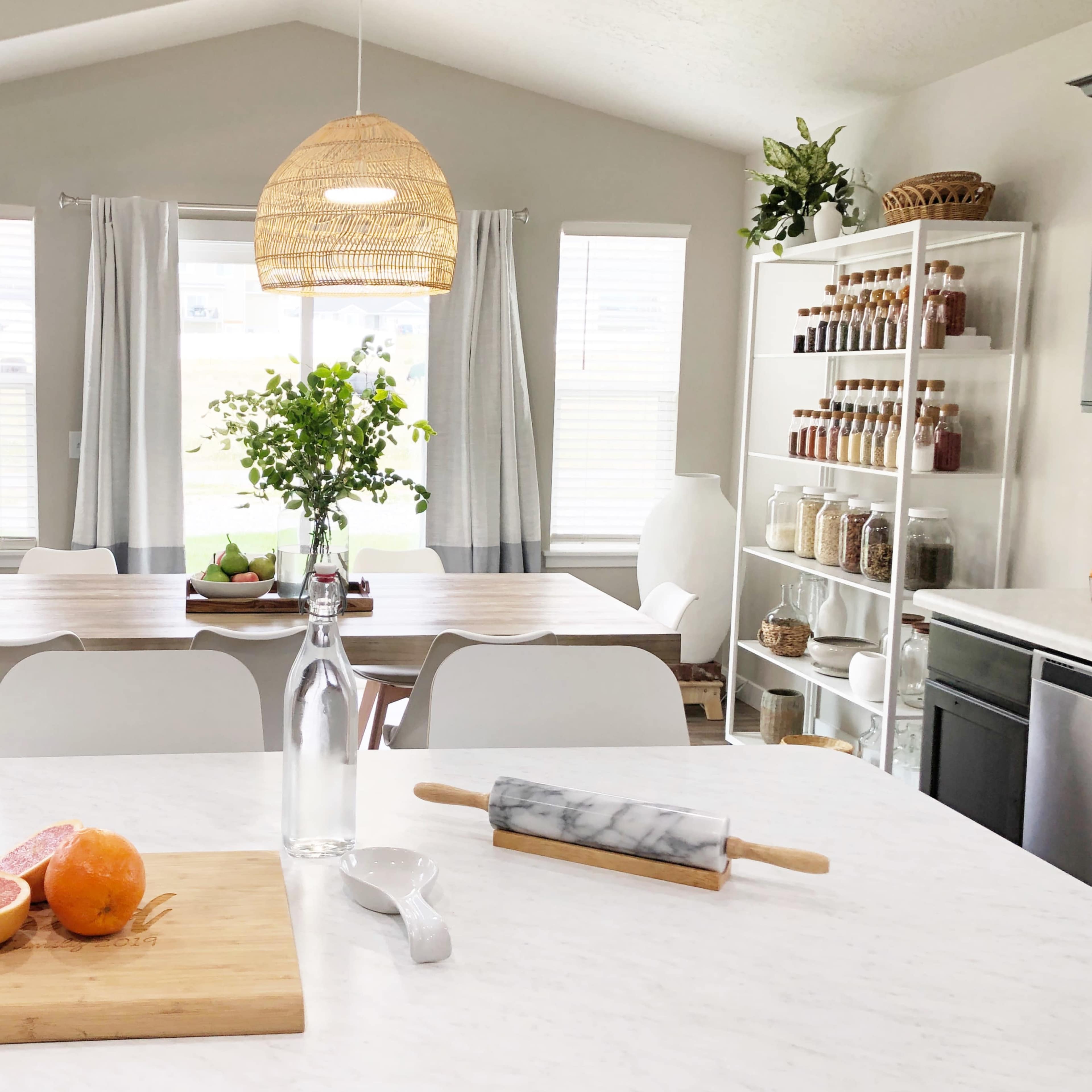 A bright and modern kitchen features a marble countertop with fruit and a rolling pin, alongside a dining area filled with a wooden table and white chairs.