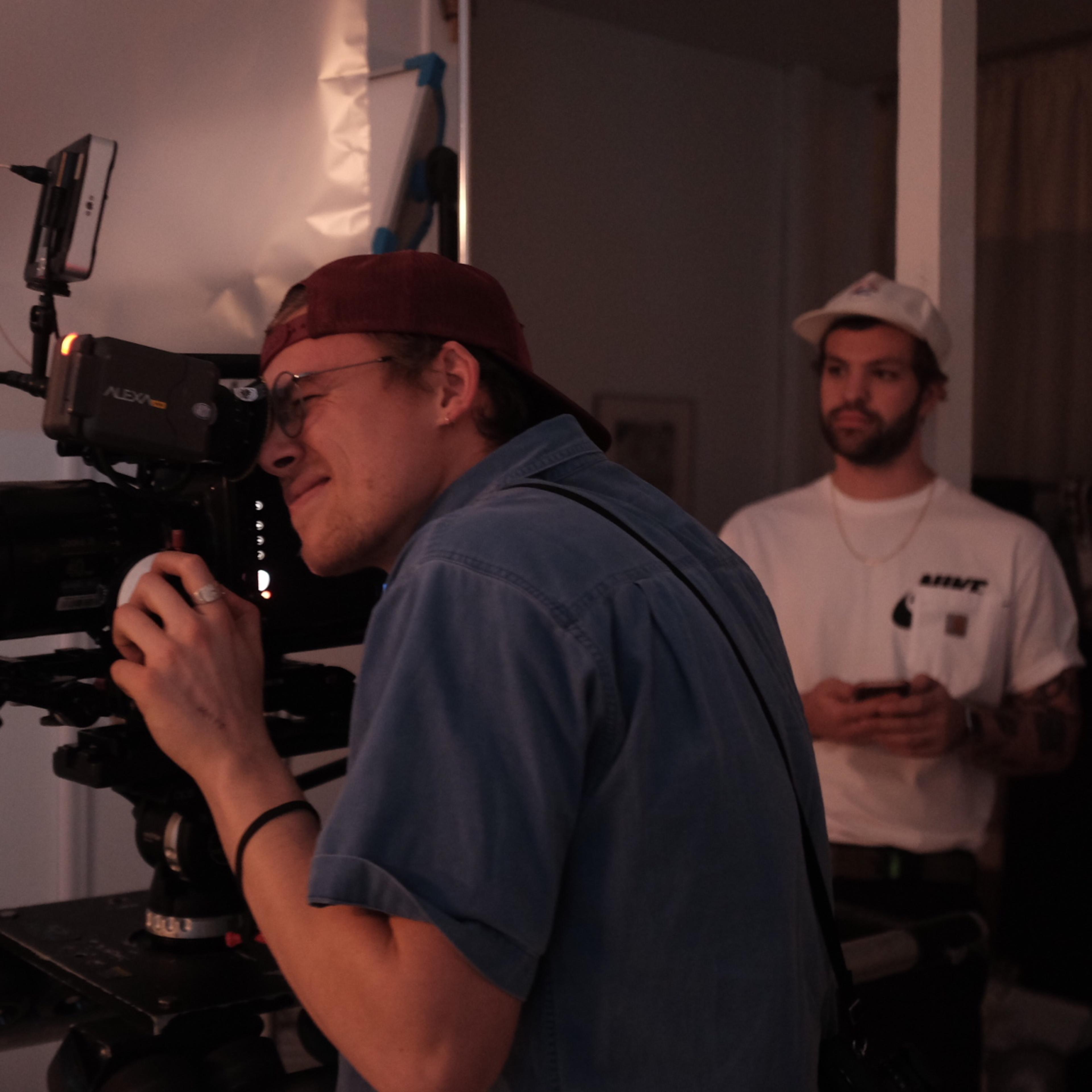 A camera operator is focused on filming with a professional camera while another person stands in the background.