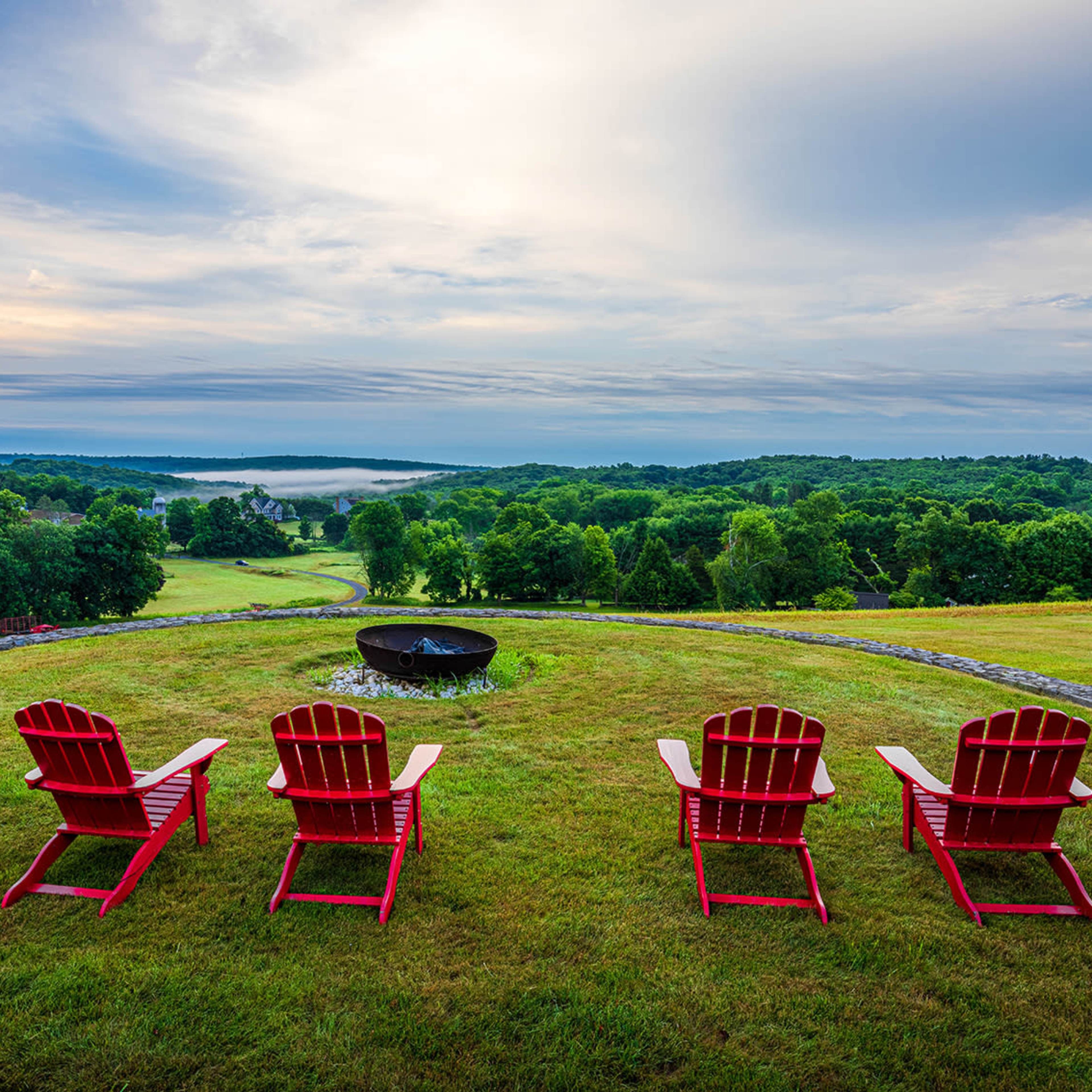 Five red Adirondack chairs are arranged in a line on a grassy hillside, facing a scenic landscape of rolling hills and a cloudy sky.