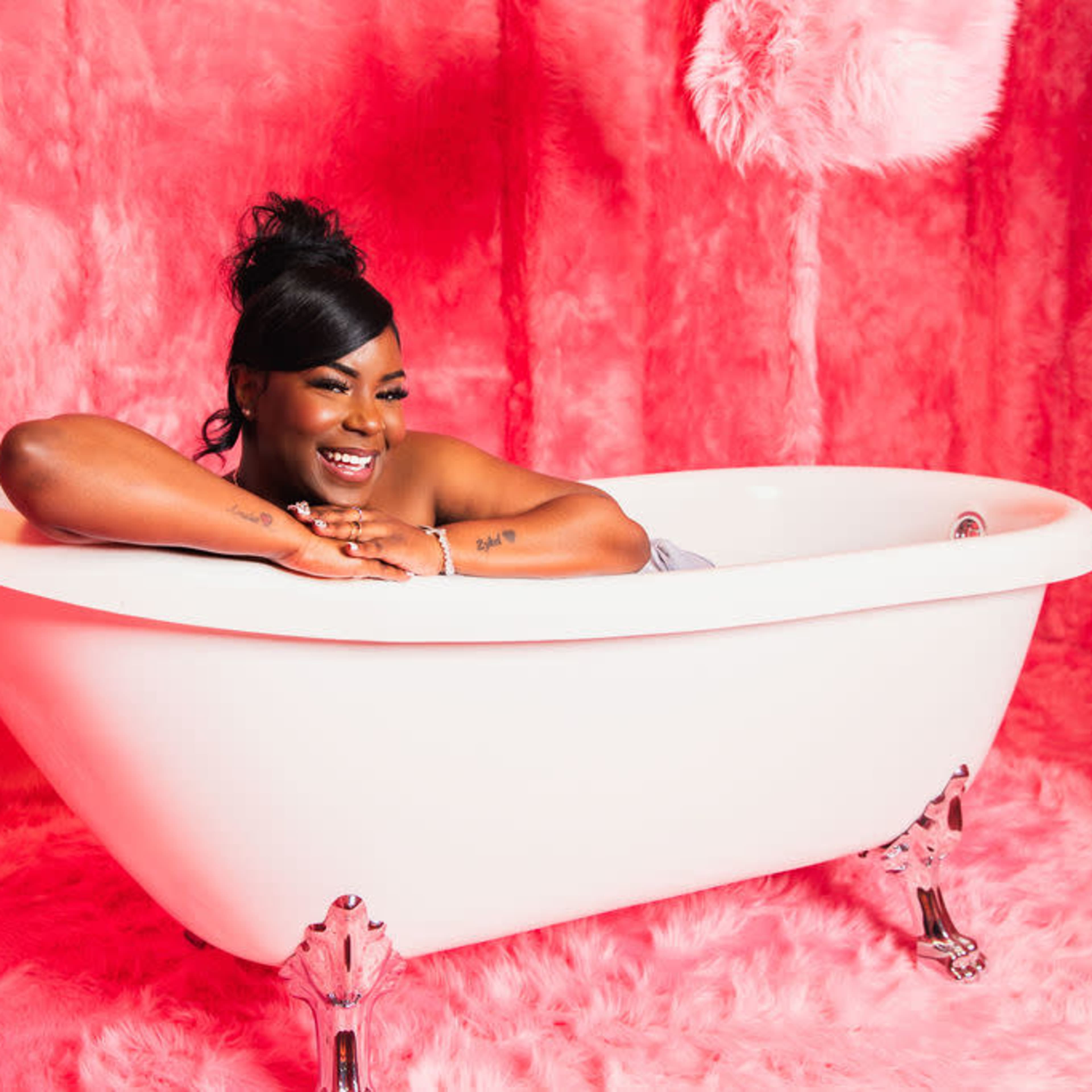A person relaxes in a white freestanding bathtub against a pink backdrop with fluffy decor.