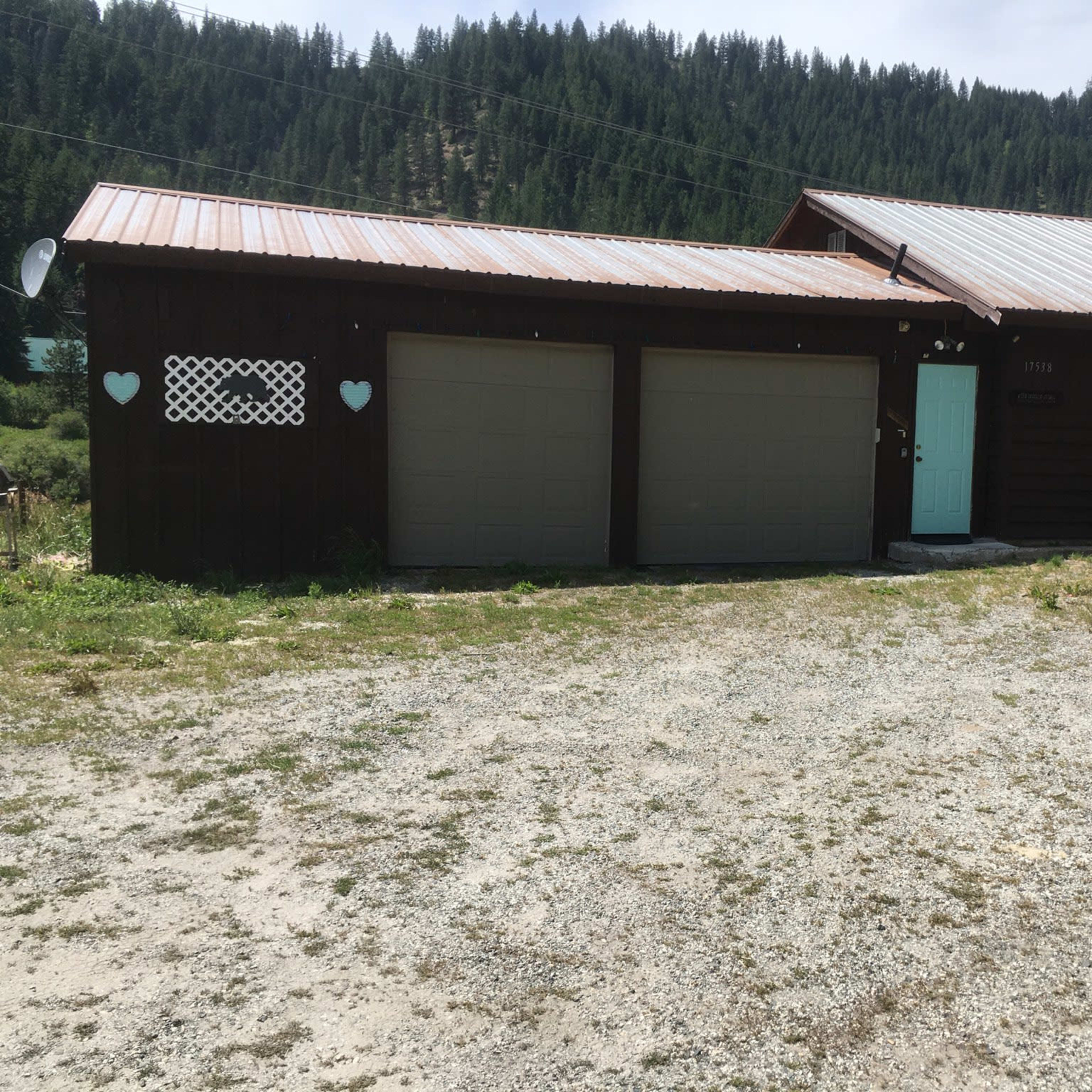 A dark wooden house with a metal roof and two garage doors, set on a gravel driveway in a mountainous area.
