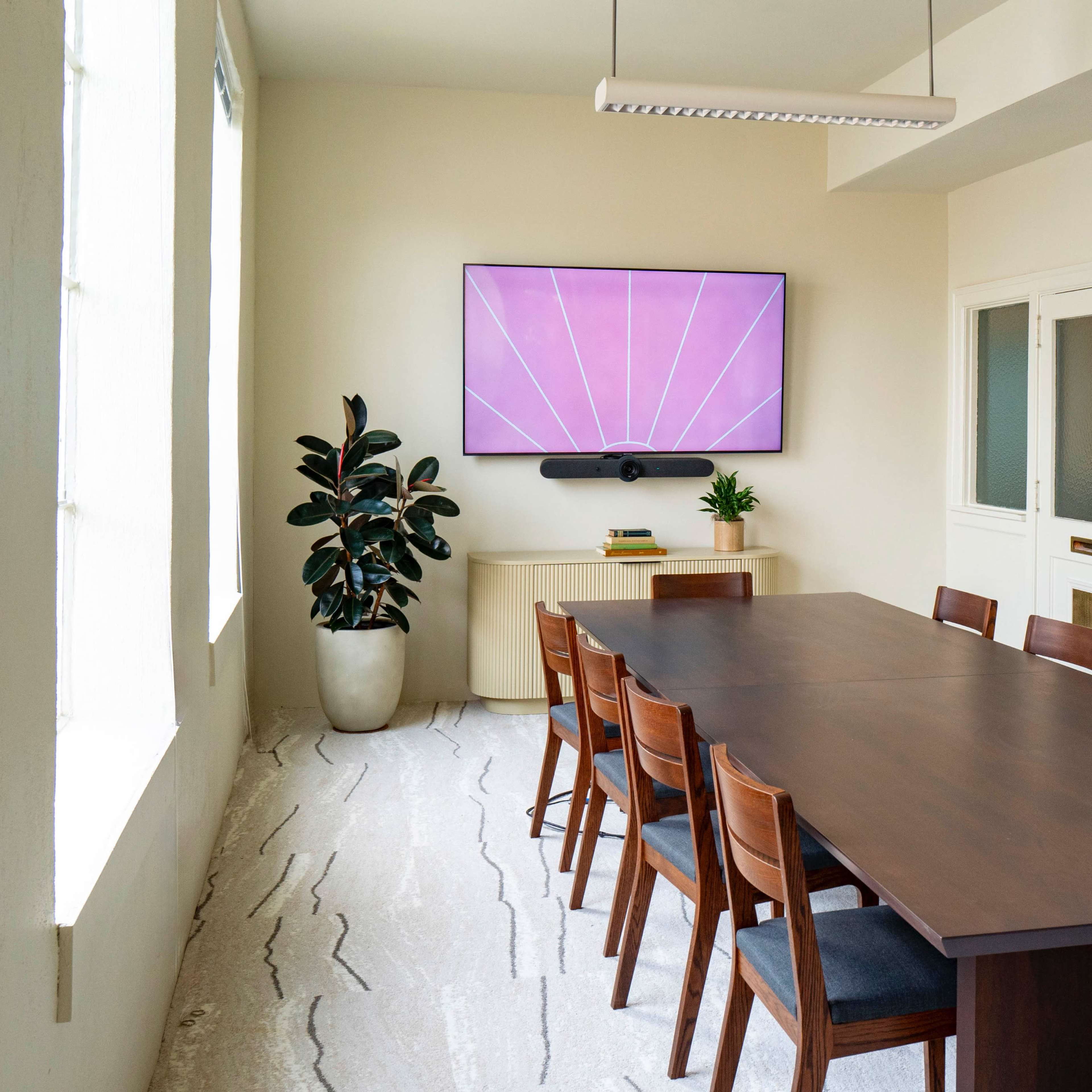 A modern conference room features a long wooden table, eight chairs, a large wall-mounted TV, and a potted plant near a window.