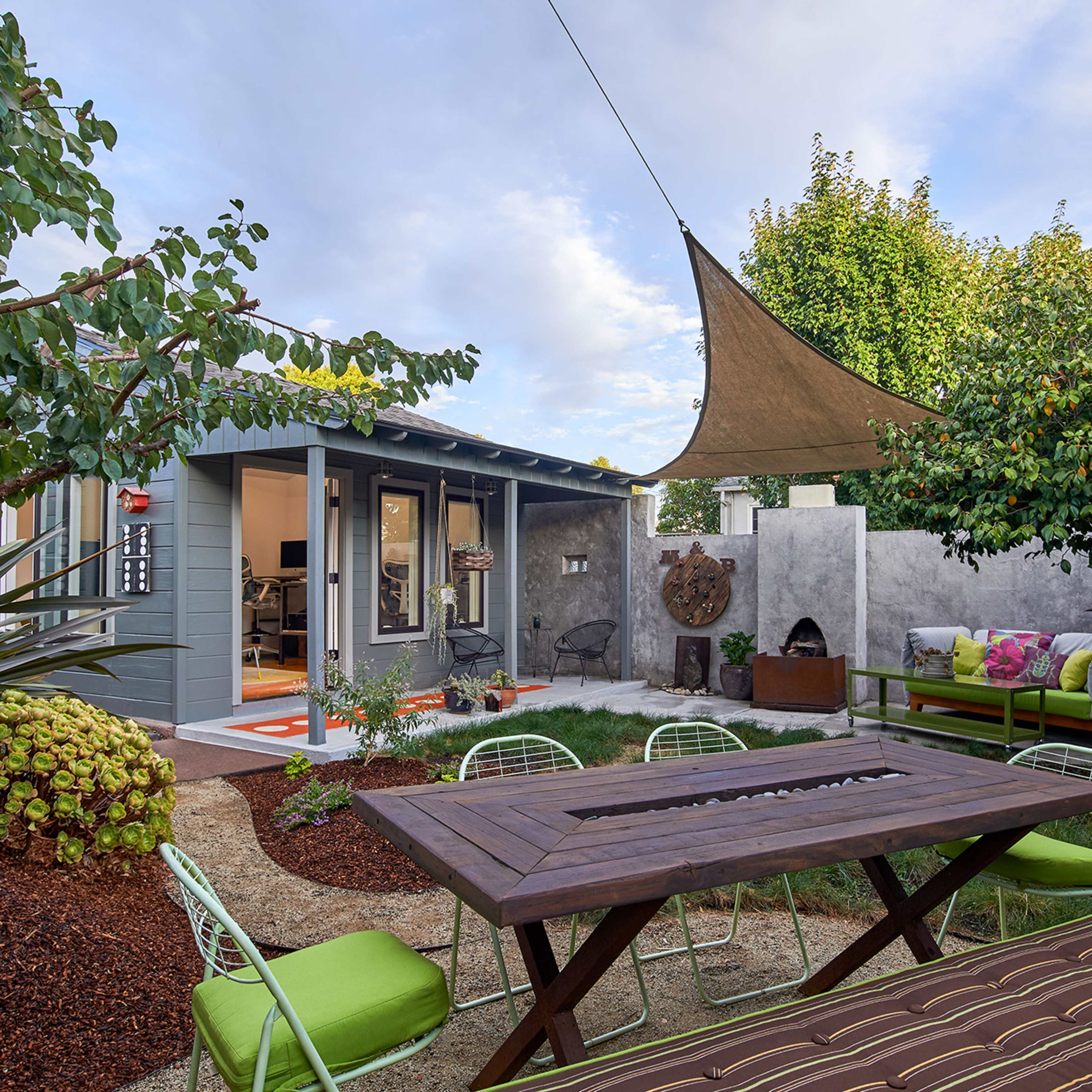 A landscaped garden featuring a wooden dining table, green chairs, and a small modern shed surrounded by trees and plants.