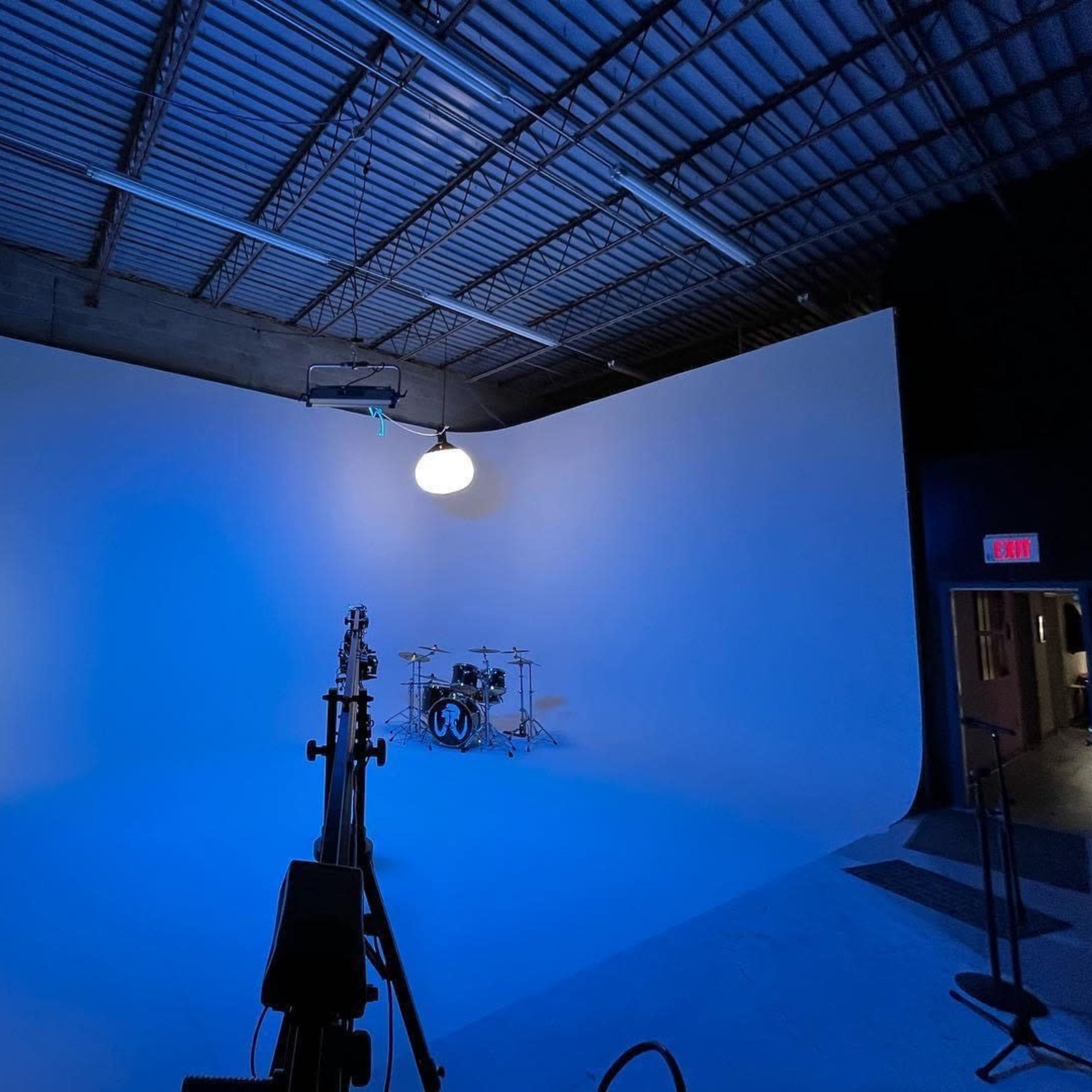 A photography studio with a seamless blue backdrop, a drum set in the background, and studio lighting equipment arranged in the foreground.