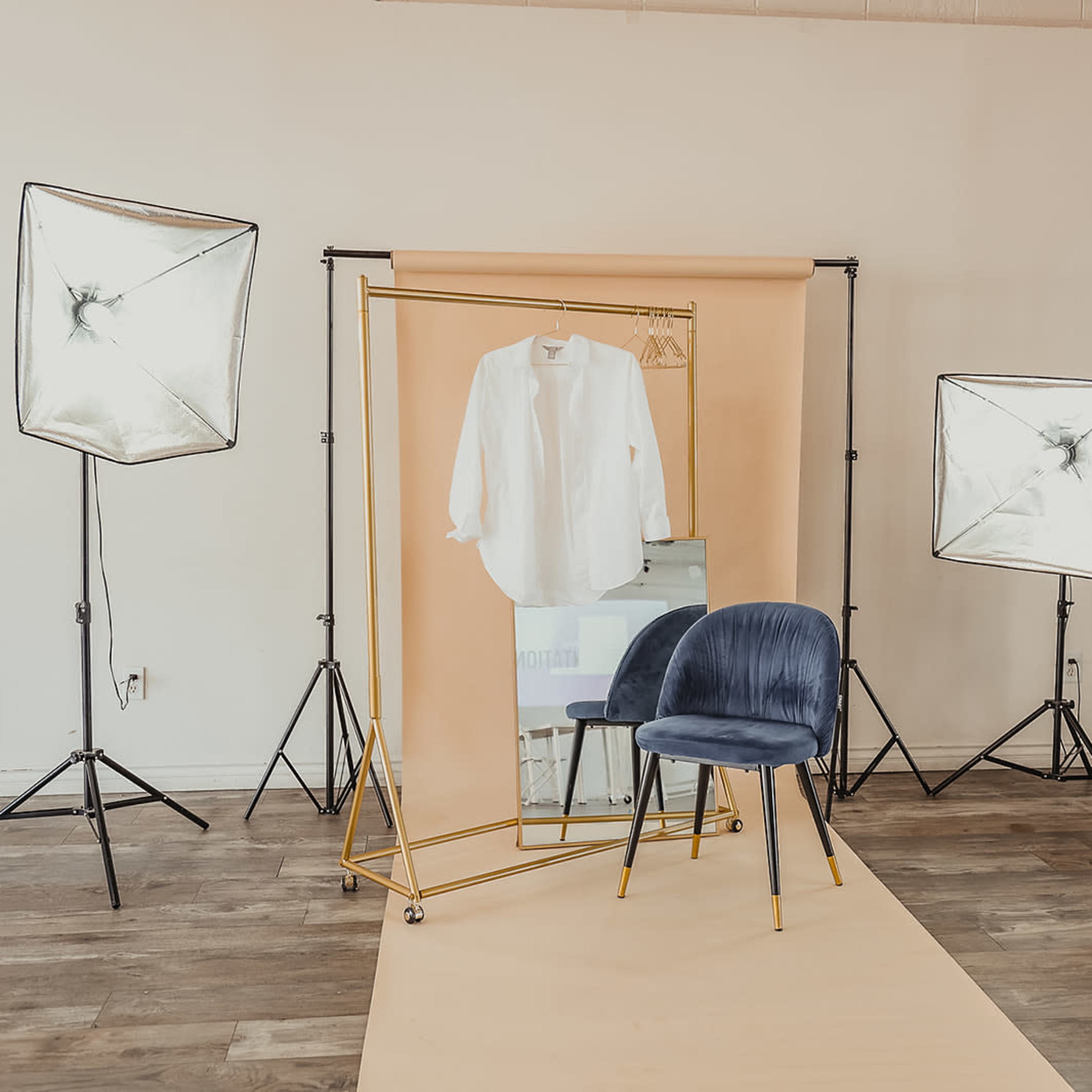 A clothing display setup with a white shirt hanging on a gold mirror surrounded by softbox lights and a blue chair against a neutral backdrop.
