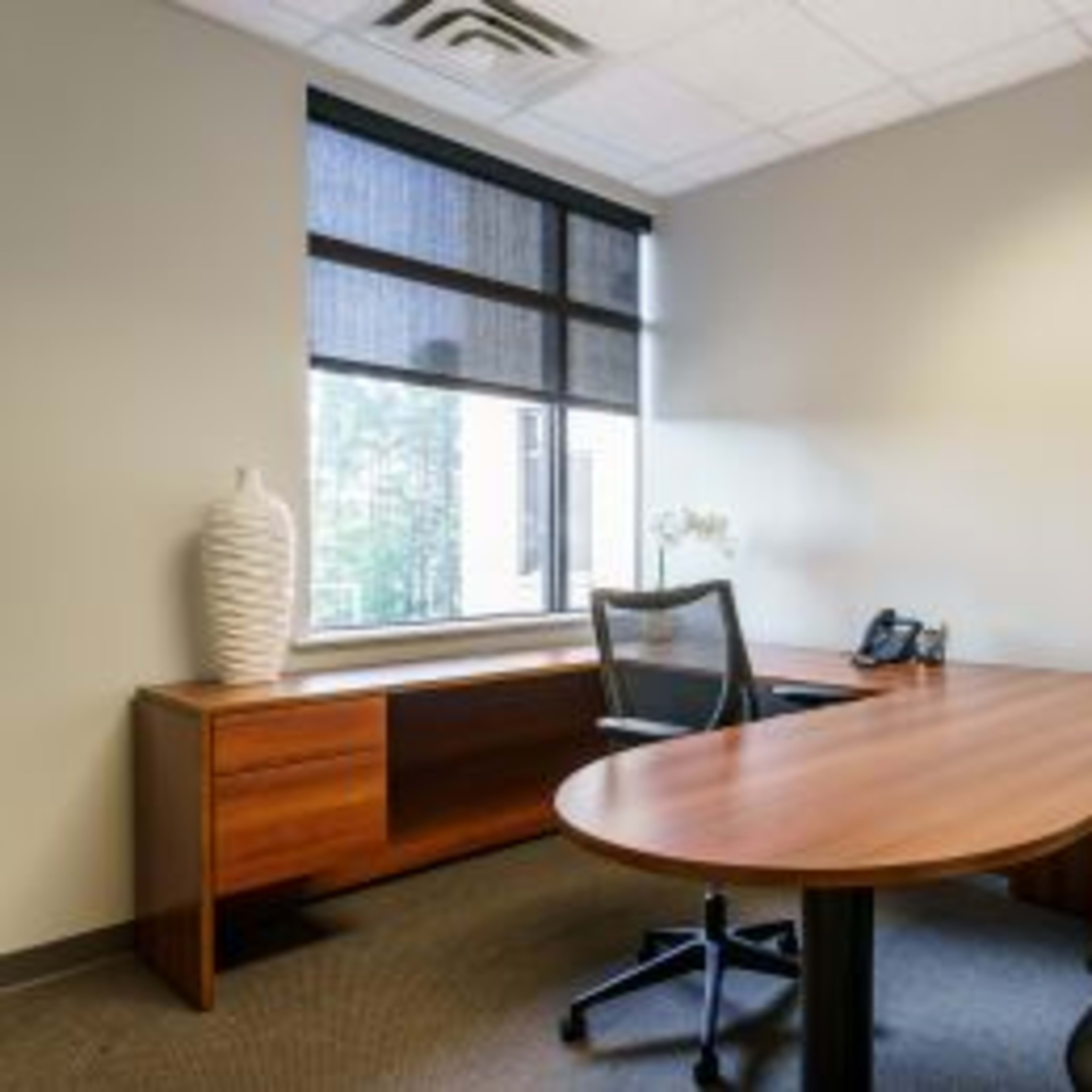 A minimalistic office space featuring a wooden desk, two black chairs, a window with blinds, and a decorative vase on a side table.