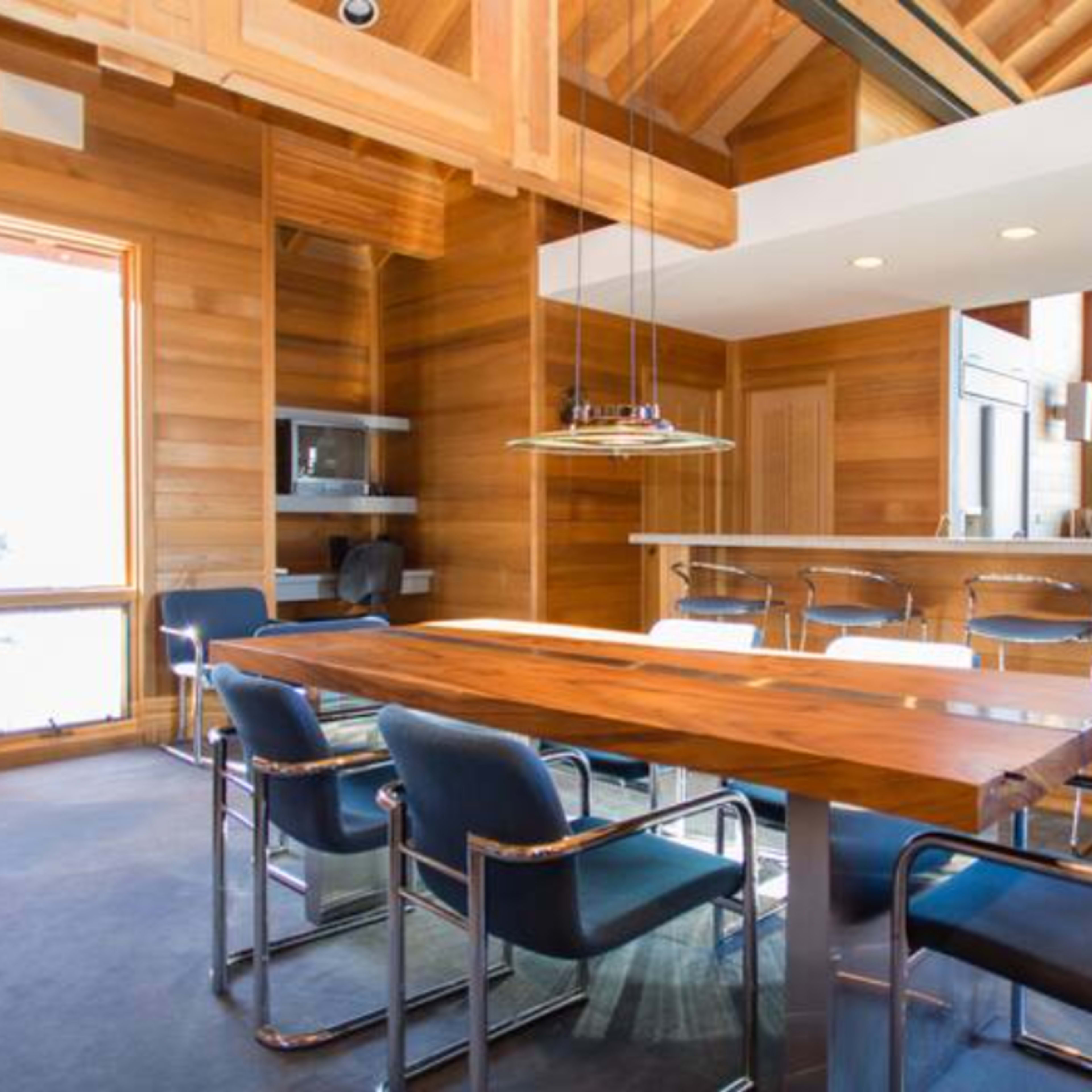 A modern dining area with a wooden table surrounded by metal-framed chairs, set in a space with wood-paneled walls and large windows.