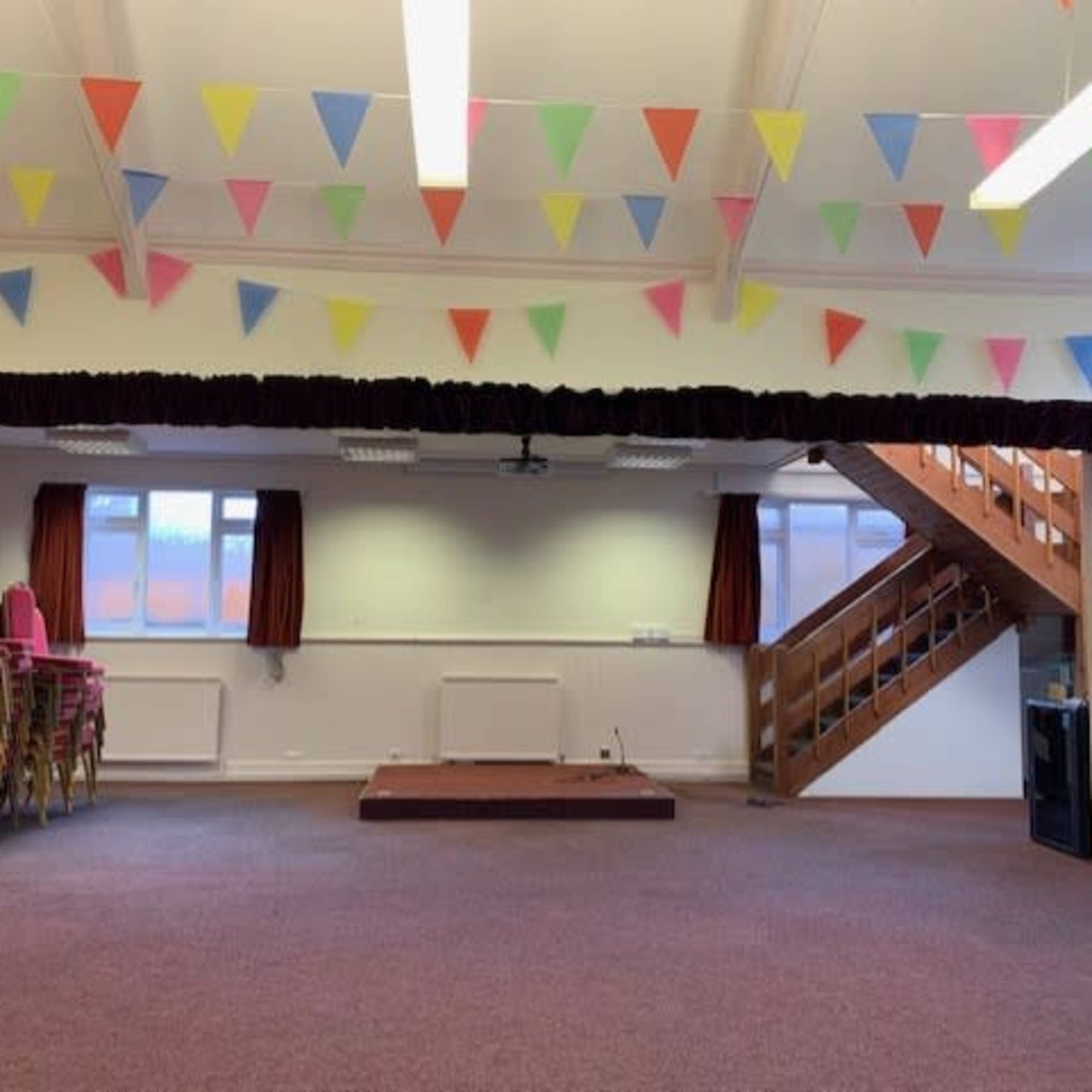 The interior of a hall features a stage framed by curtains, with colorful bunting hanging from the ceiling and chairs stacked against the wall.