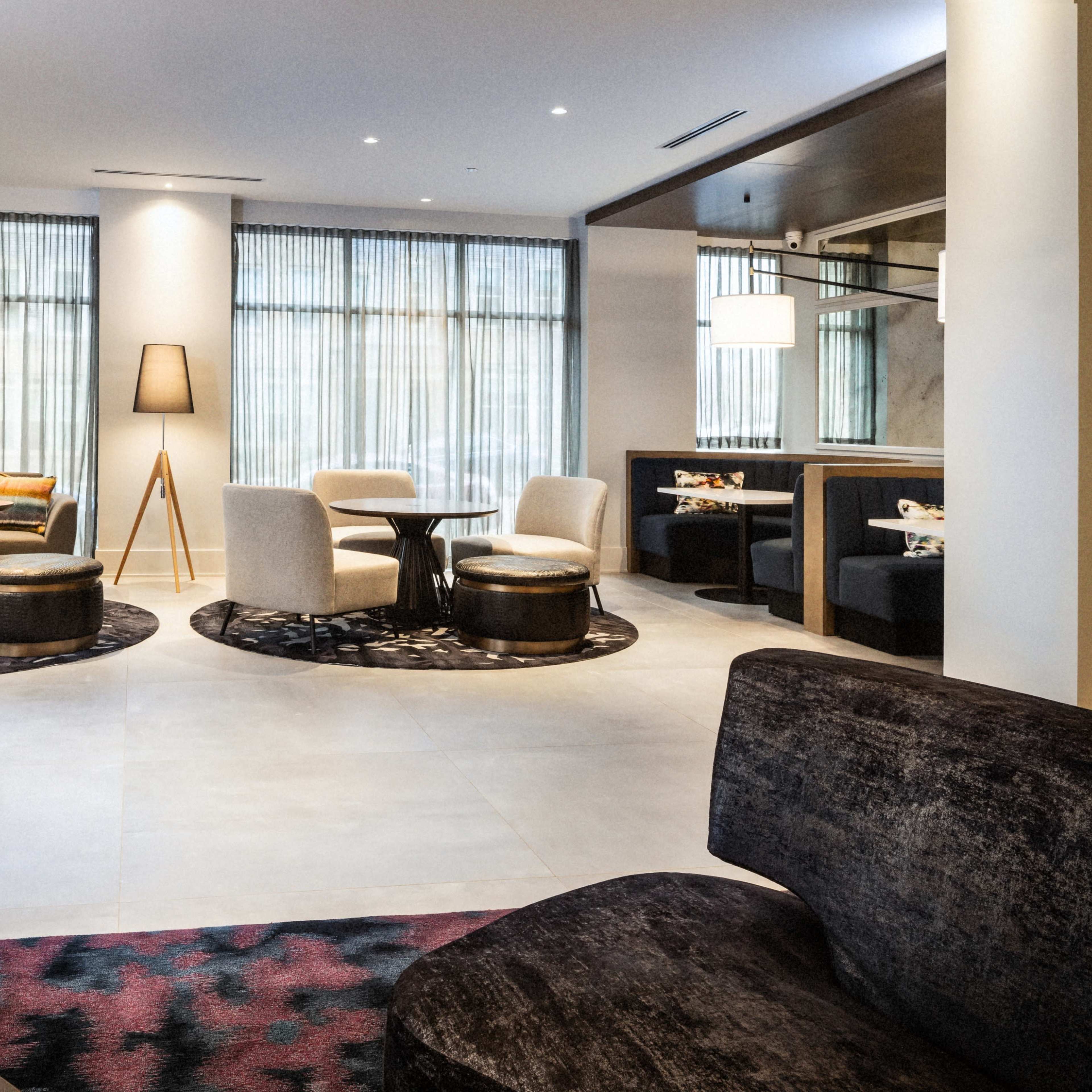 A contemporary lounge area featuring various seating arrangements, including chairs and tables, set against large windows with sheer curtains.