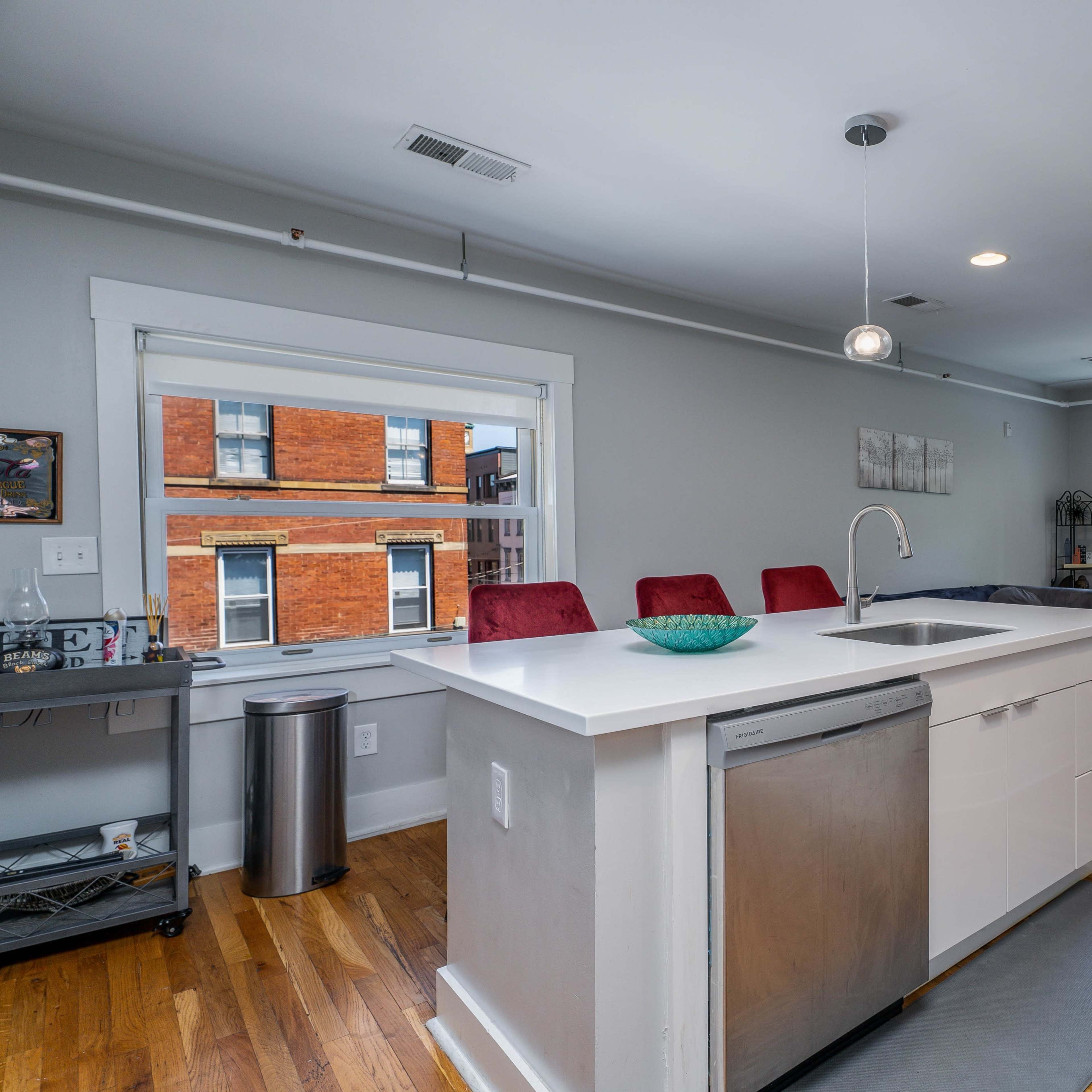 A modern kitchen with a white island and stainless steel appliances, adjacent to a living area featuring a television and red seating.
