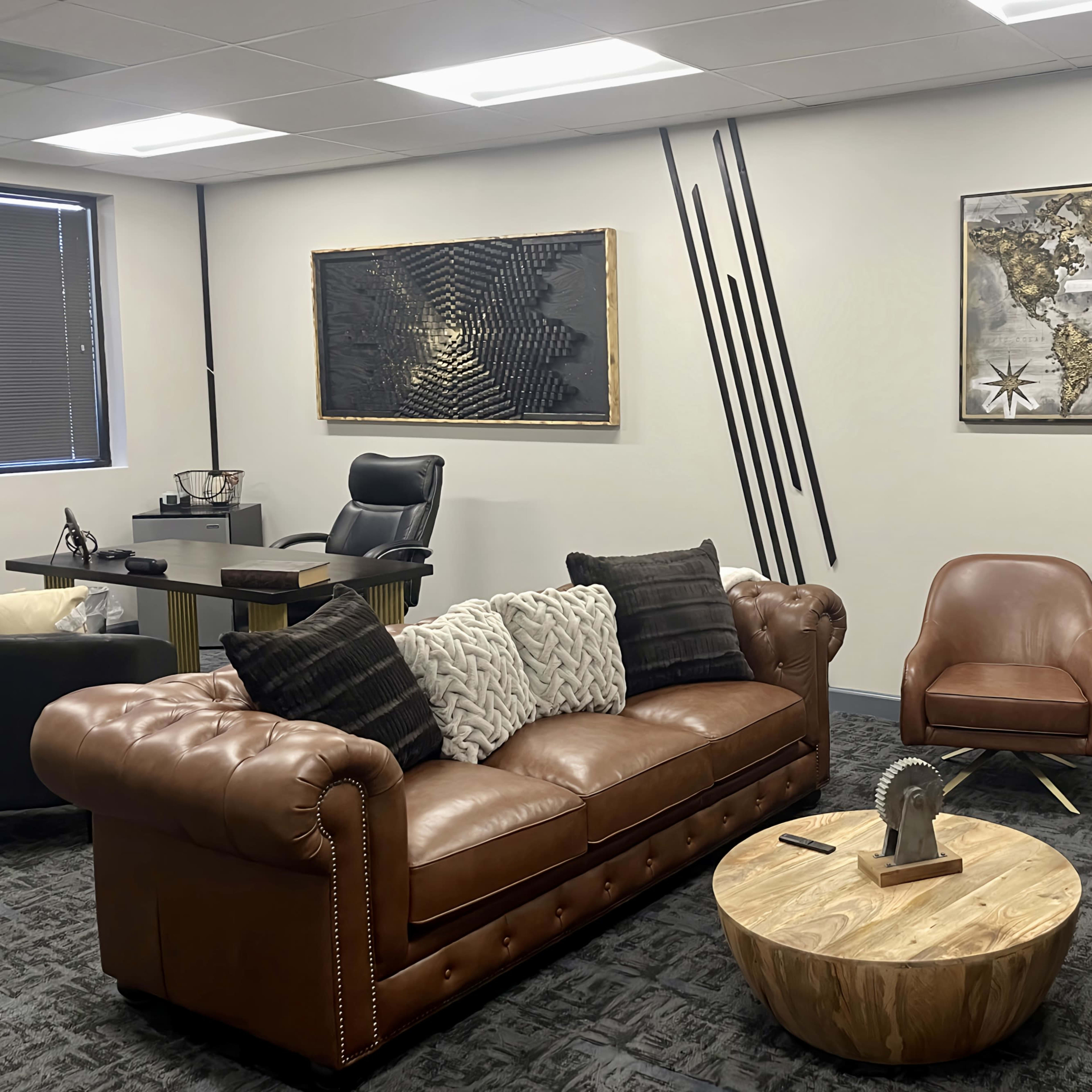 A modern office space featuring a brown leather sofa with decorative pillows, a round wooden coffee table, two stylish chairs, and wall art including a world map and abstract designs.