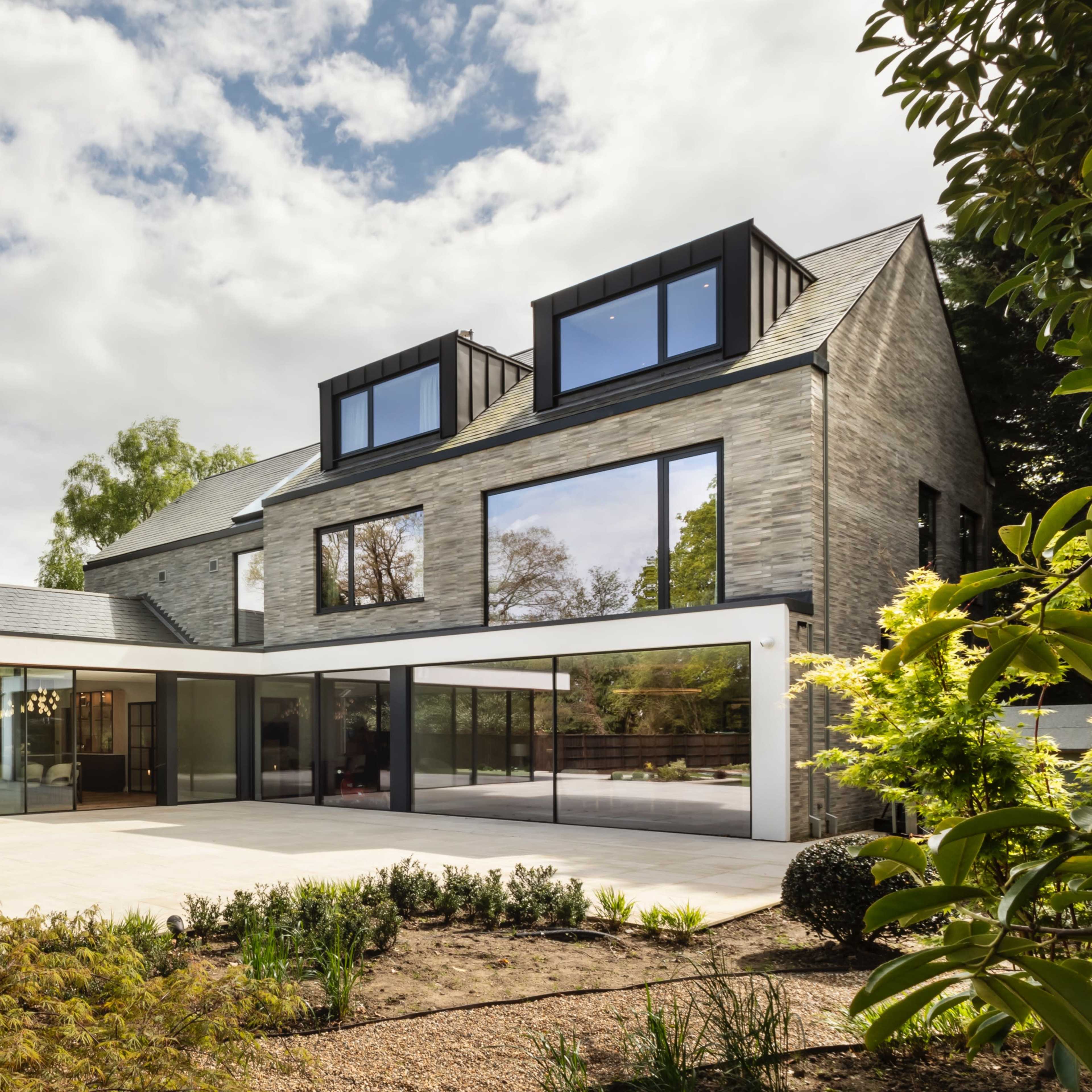 A modern two-story home with large glass windows, a flat roof section, and a stone exterior surrounded by landscaped greenery.